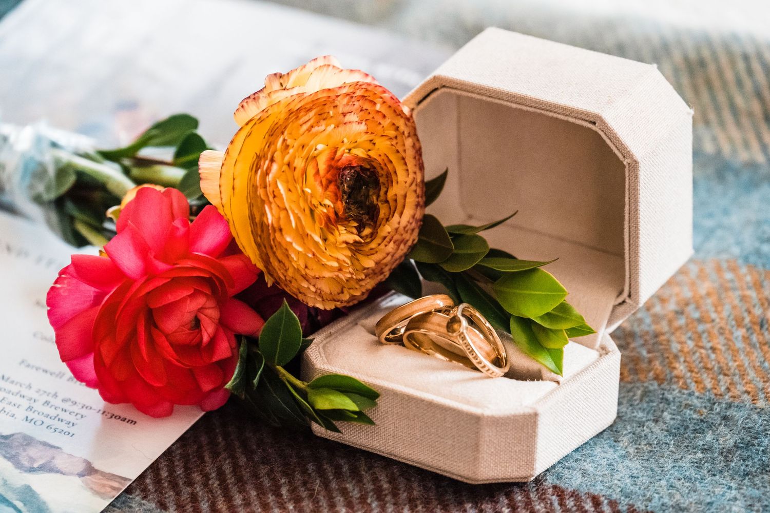 Orange ranunculus and red rose flowers with wedding rings in an open white box.