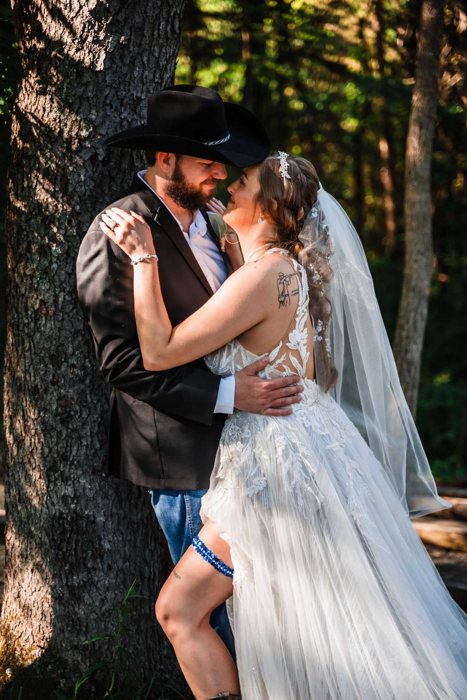 A romantic wedding couple embraces against a tree in the forest, with the bride wearing a flowing white gown and veil.