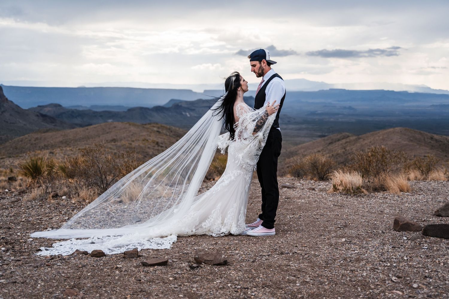 A couple in wedding attire stands atop a mountain with a flowing veil and dramatic mountain landscape behind them.