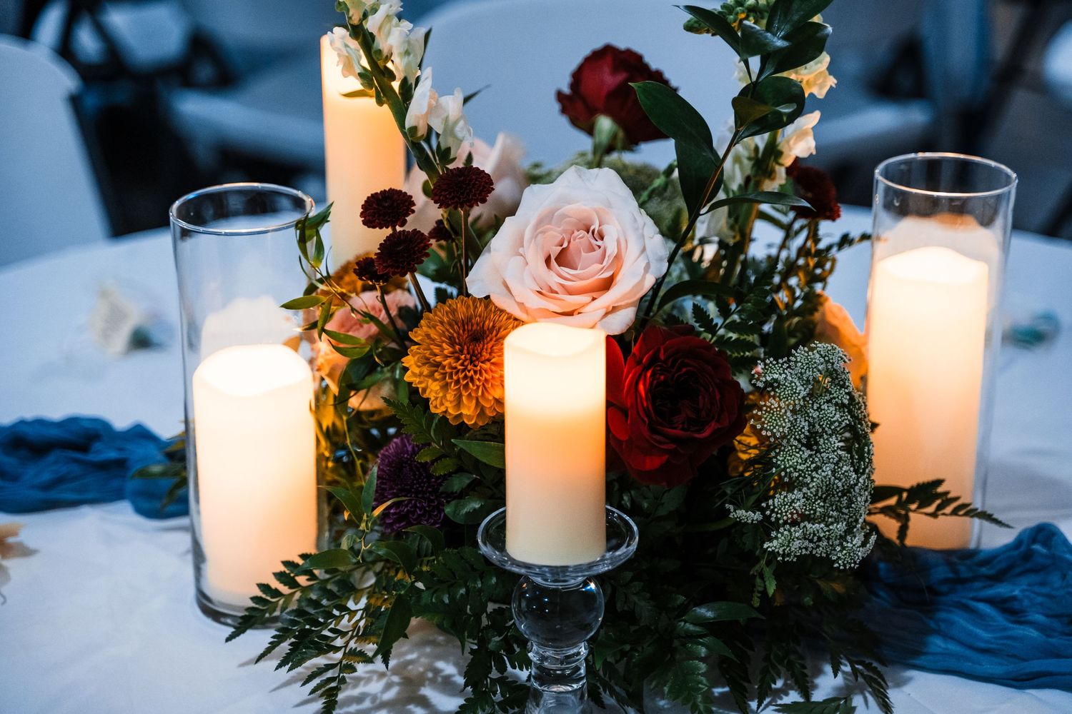 Elegant wedding centerpiece with roses, candles and greenery on blue tablecloth.