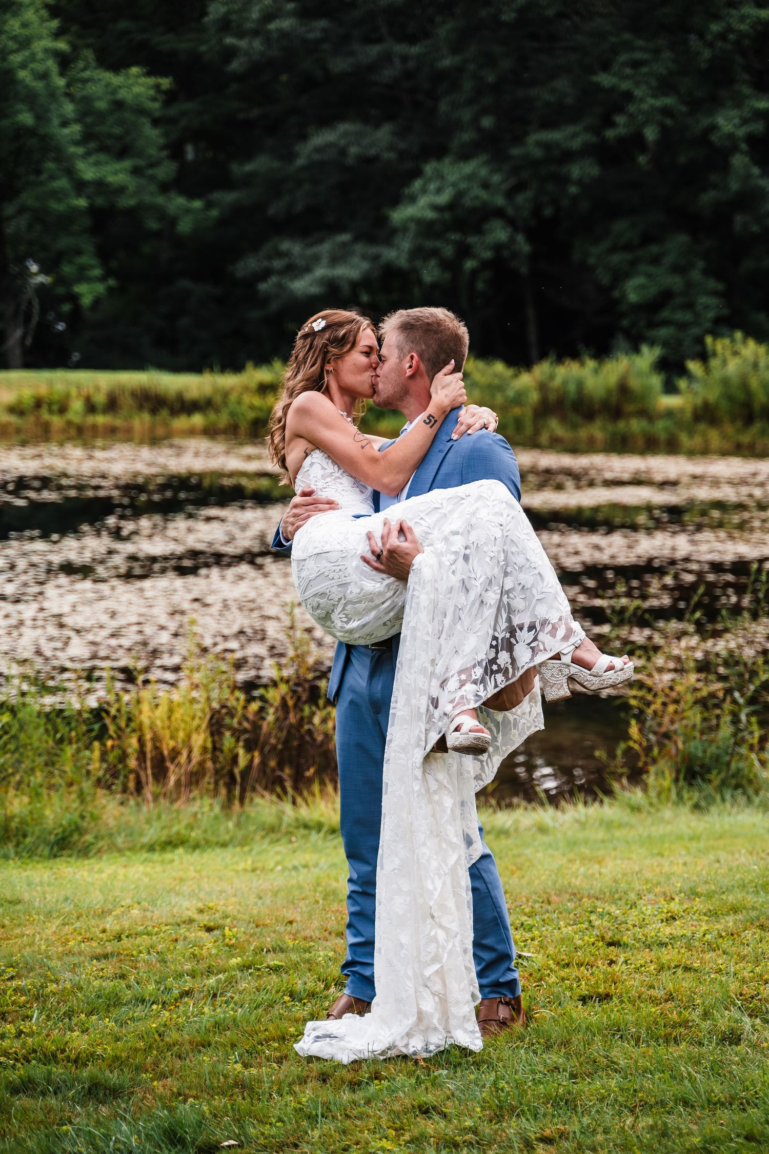 A romantic lift pose during wedding photos beside a scenic pond with trees in the background.