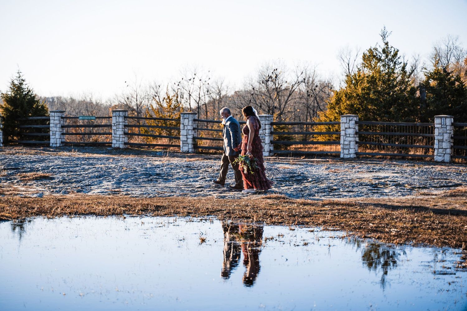 Couple's reflection in puddle with rustic wooden fence and winter sky backdrop.