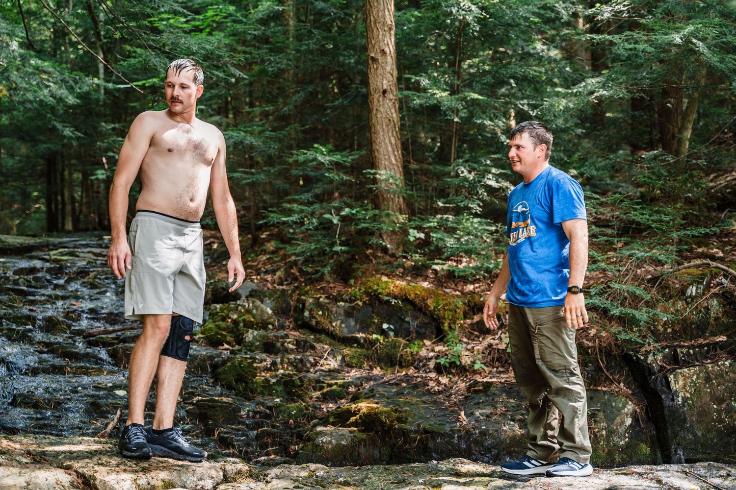 Two hikers stand on rocks near a forest stream on a sunny summer day.