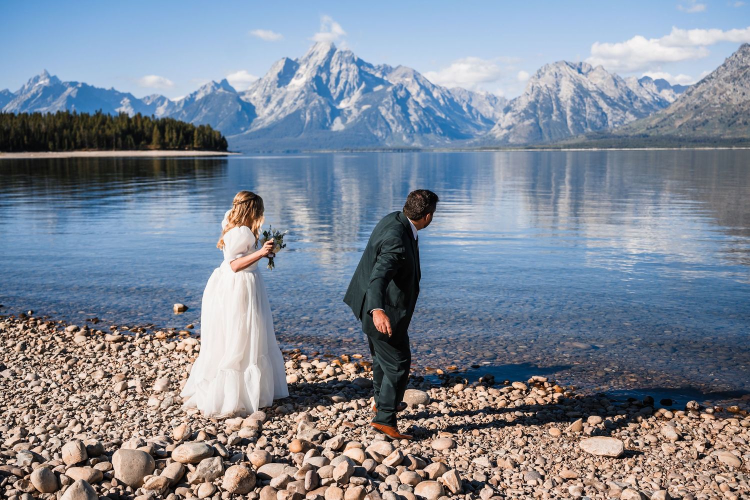 Two people in wedding attire stand on a rocky shoreline with snow-capped mountains and pristine lake waters in the background.