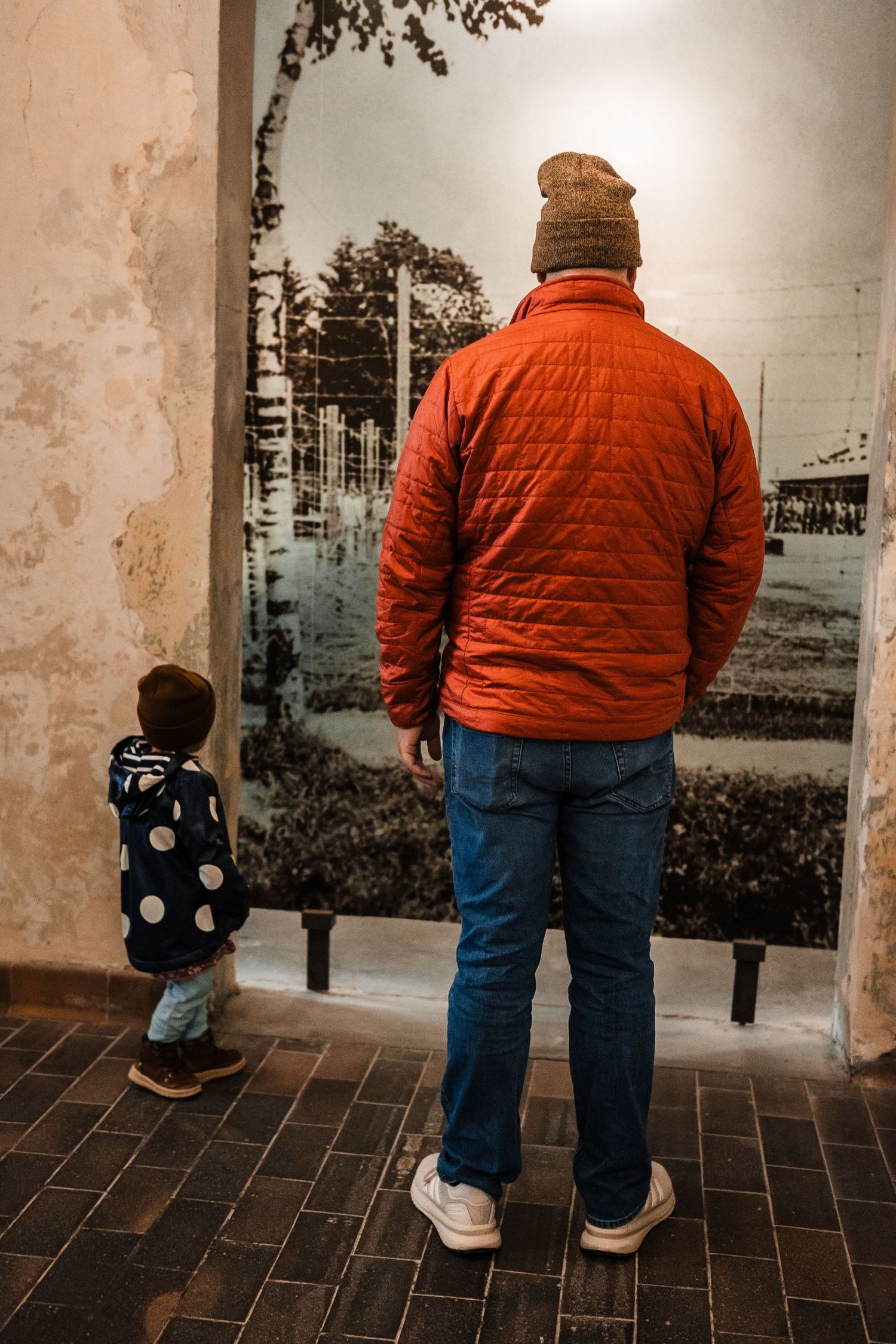A person in an orange jacket and jeans kneels down in front of a large wall mural in a gallery setting.