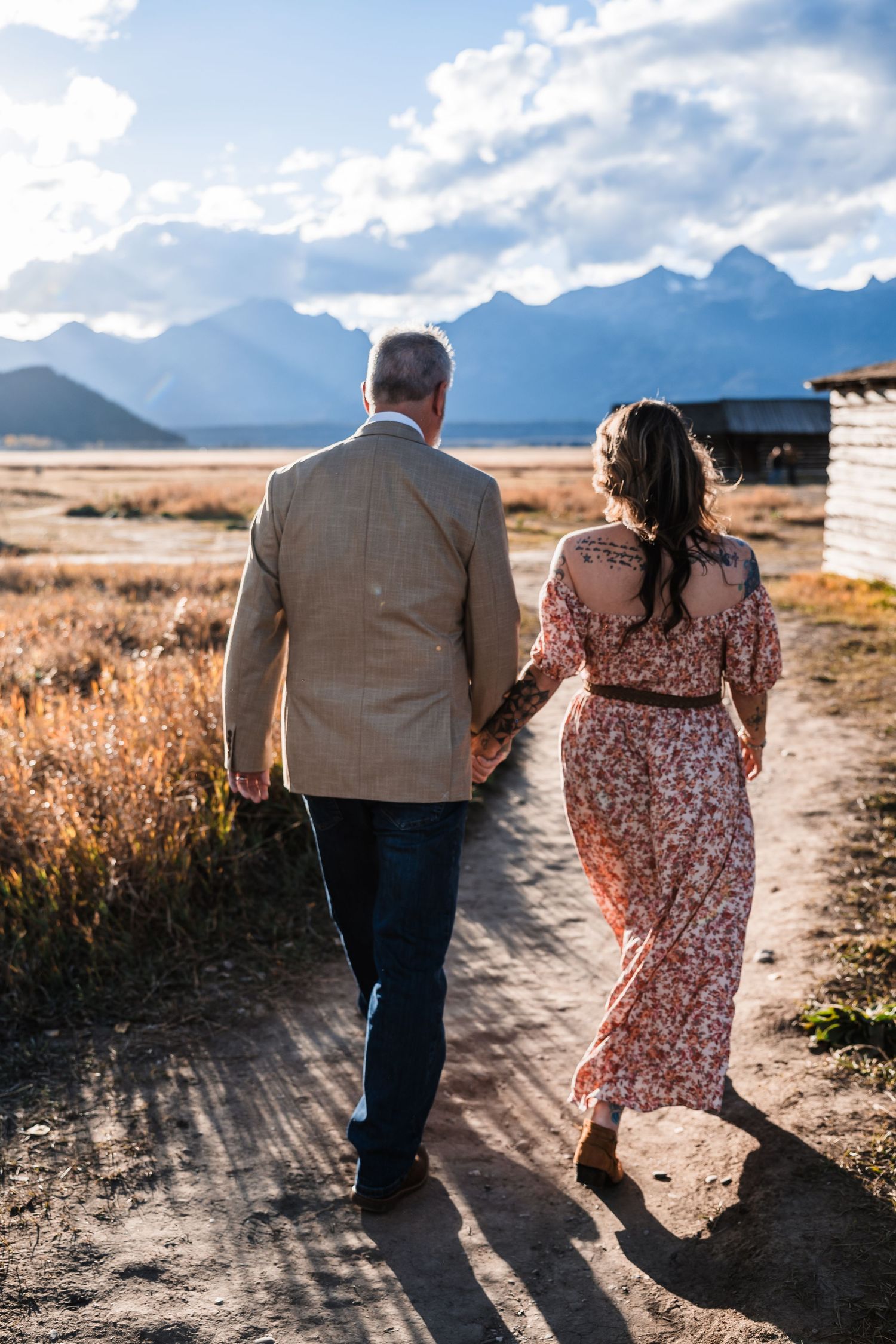 A couple walks hand in hand along a dirt path with mountains and cloudy skies in the background.