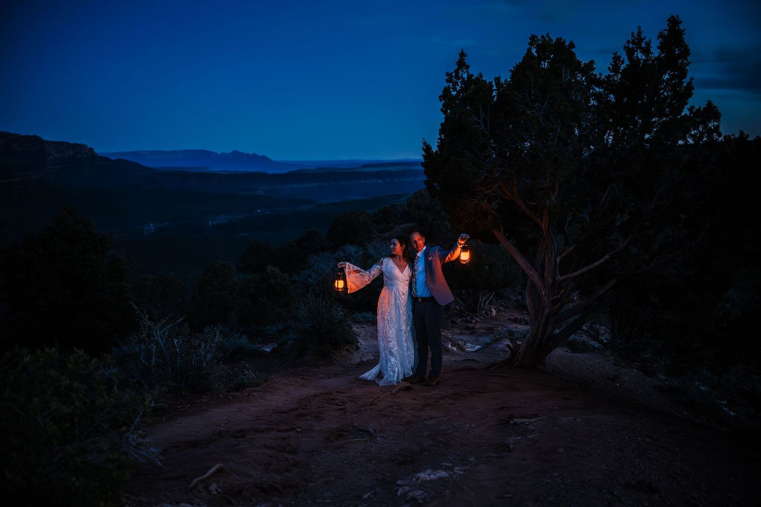Bride and groom hold glowing lanterns at dusk on scenic canyon overlook at night.