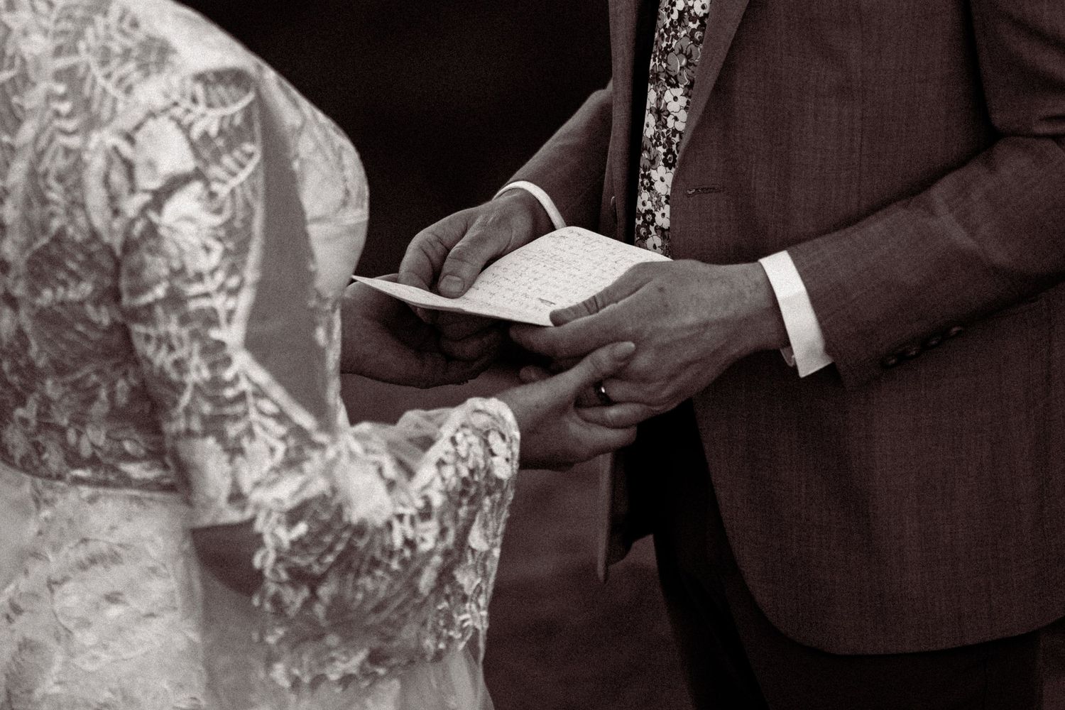 Black and white photo of groom reading wedding vows from a book while holding hands with bride in lace dress.