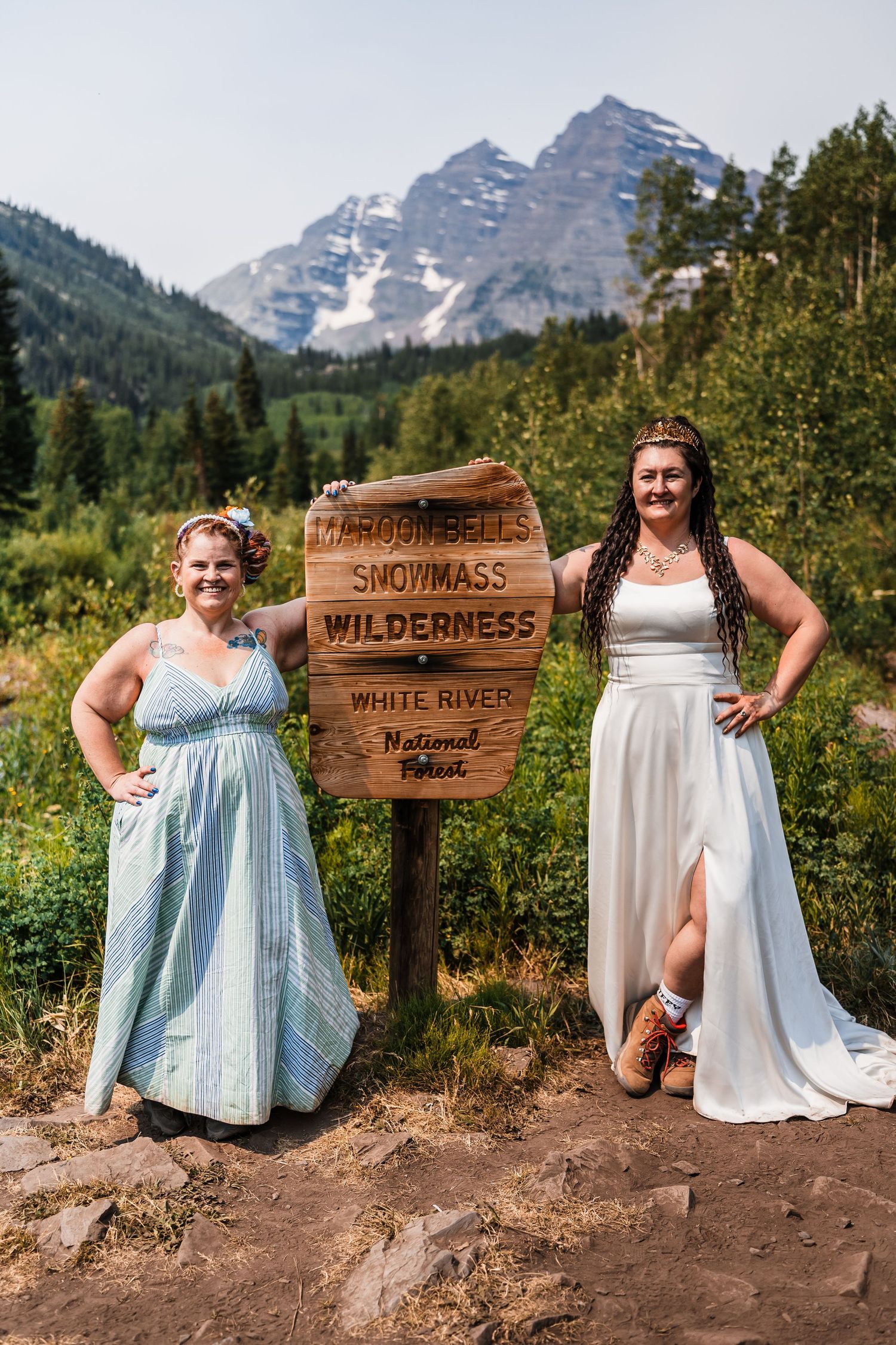 Two women in white dresses pose at Maroon Bells Snowmass Wilderness sign in White River National Forest Colorado.
