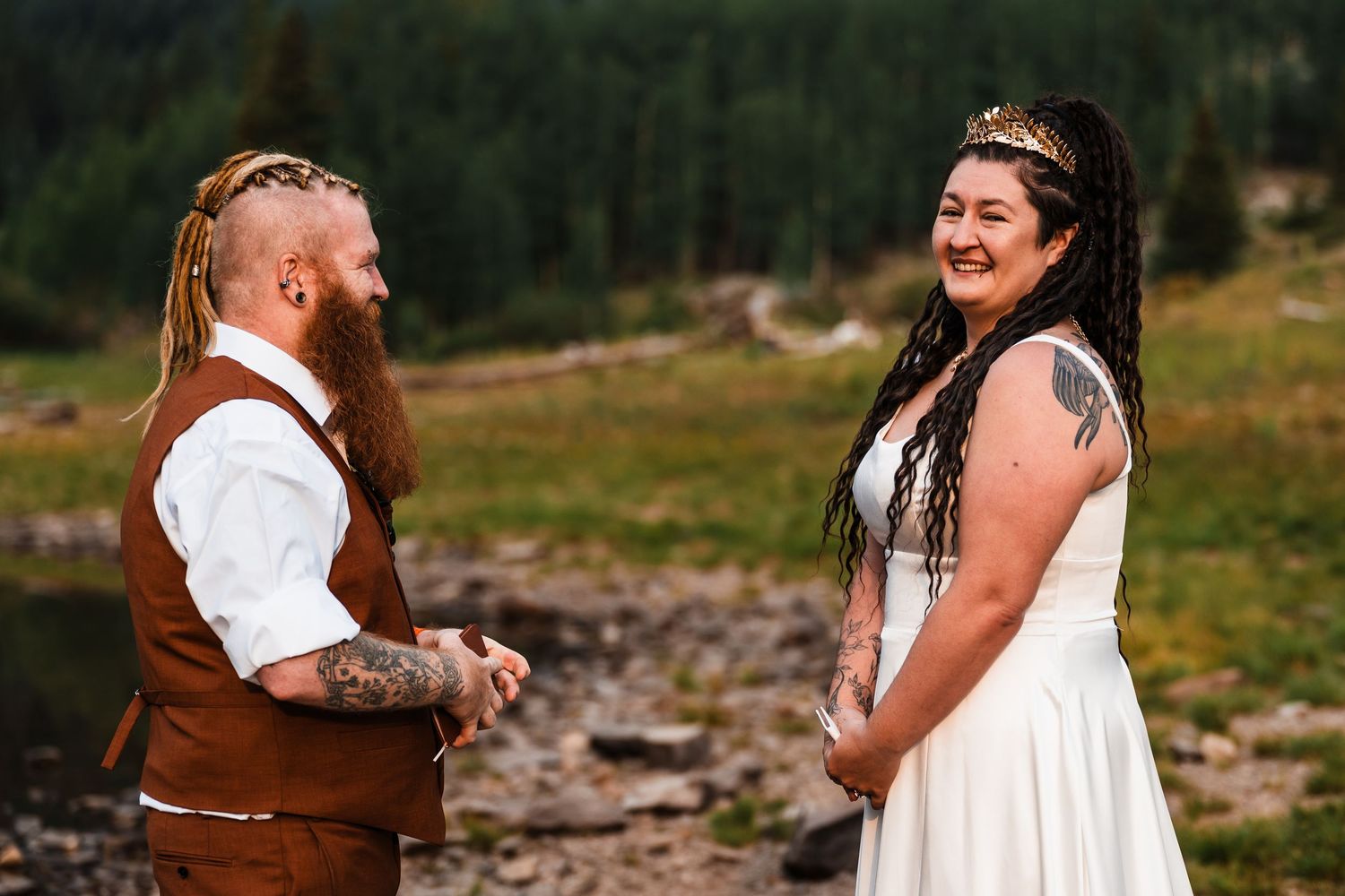 Tattooed bride in white dress laughs joyfully while holding hands with red-bearded groom during outdoor mountain elopement.