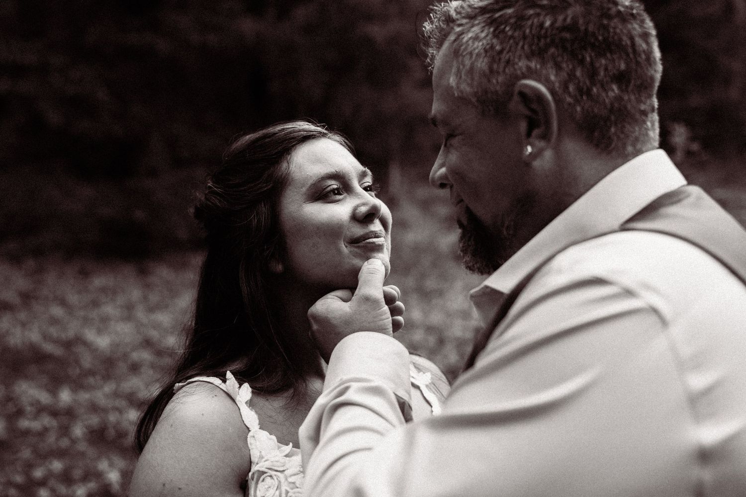 Groom tenderly touches bride's face in intimate black and white wedding portrait outdoors.