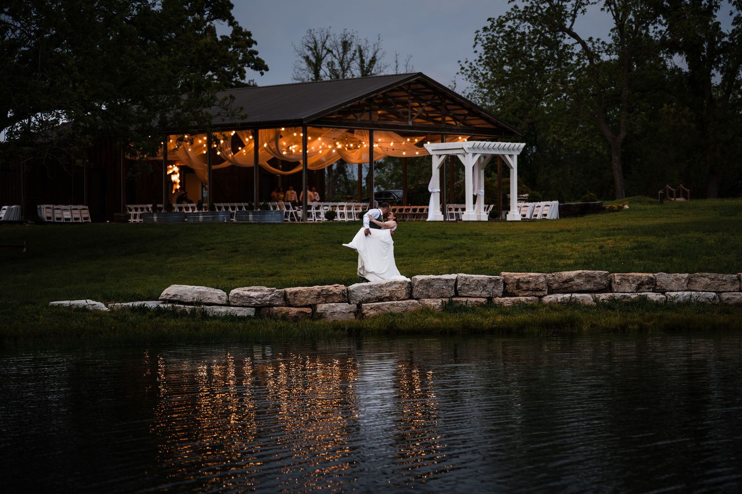 Bride and groom sit by a pond at dusk with lit wedding venue reflecting in water.