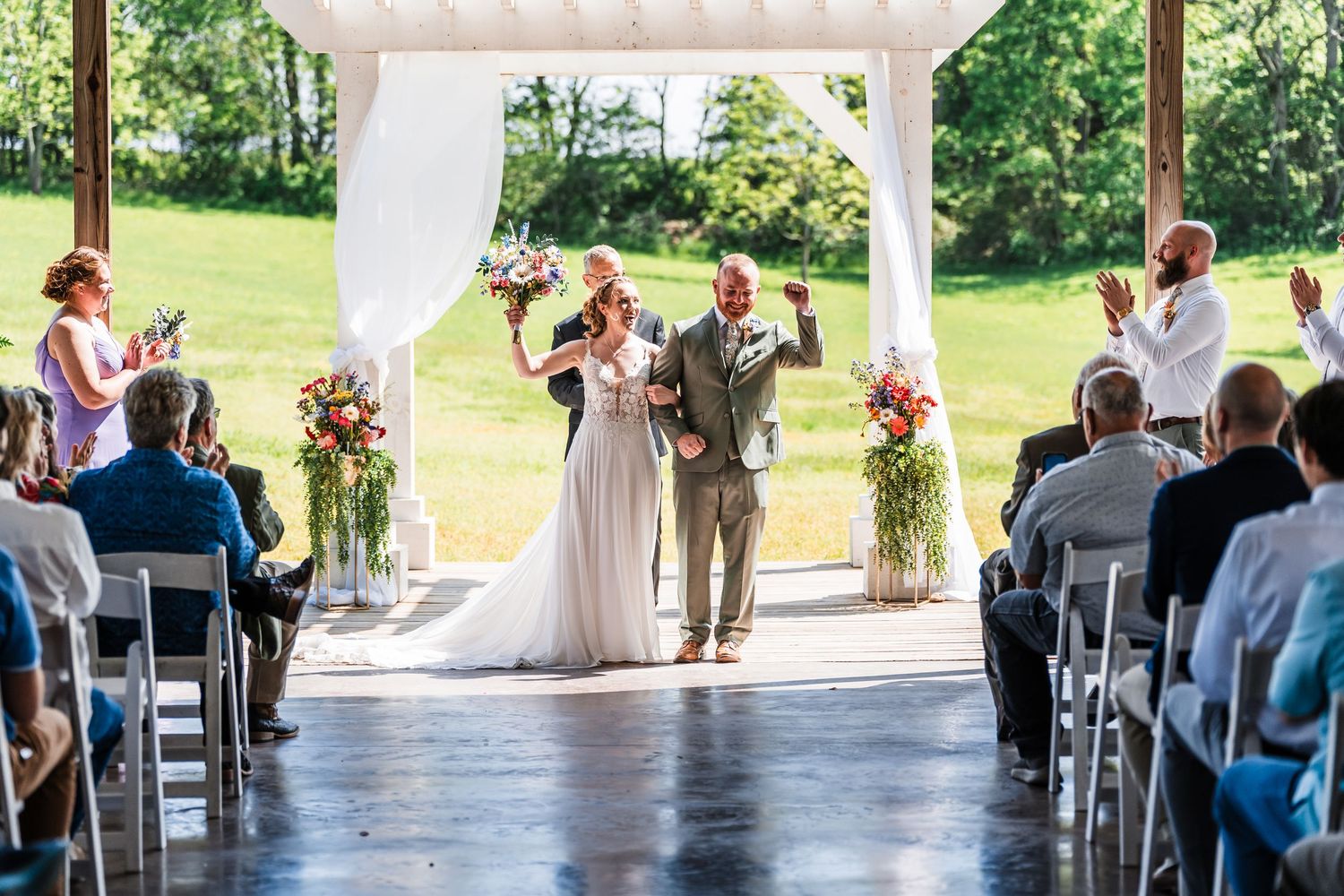 Newlyweds celebrate joyfully as they are introduced under a white wedding arch with applauding guests on a sunny day.