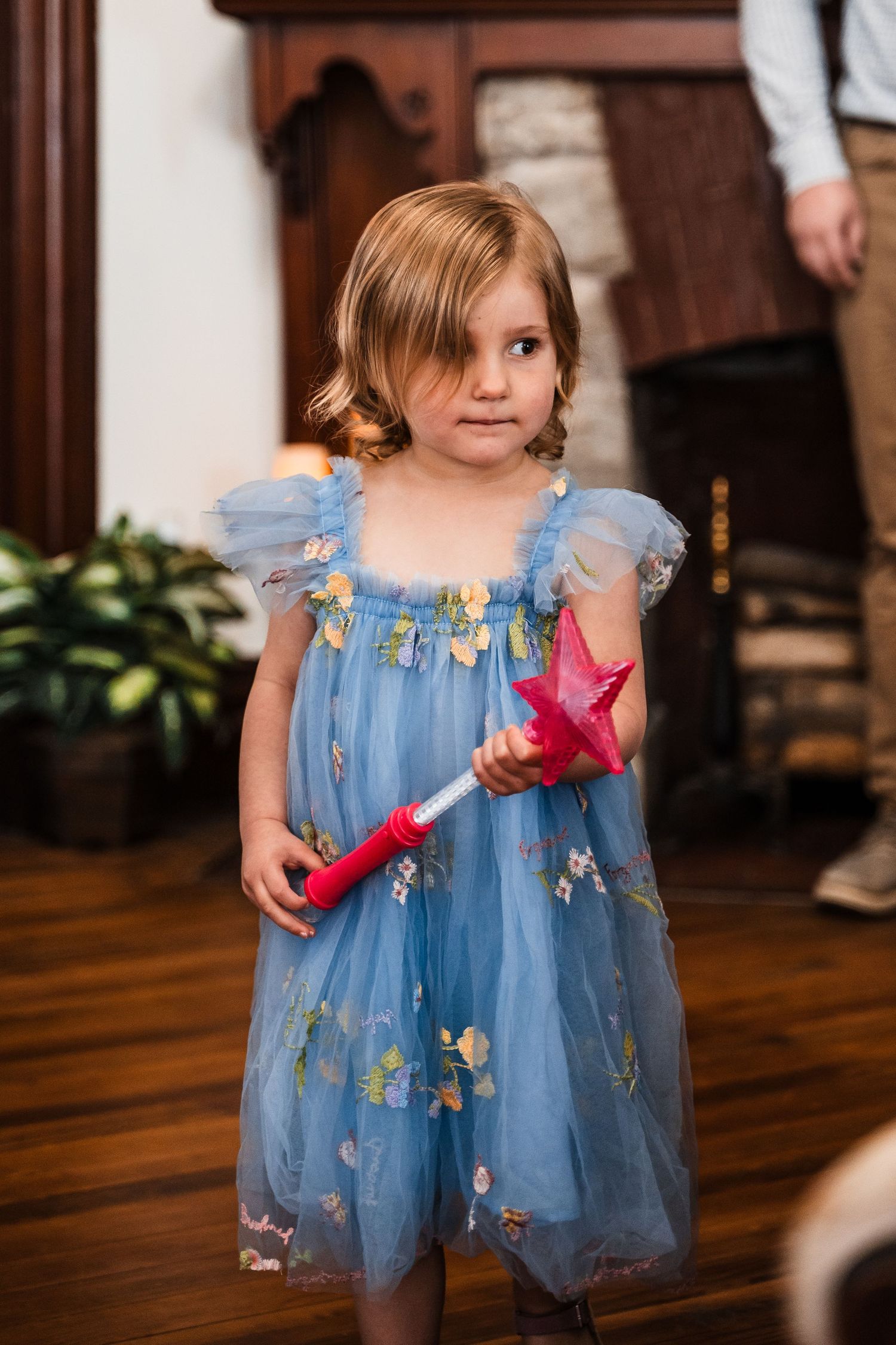 Young girl in blue floral tulle dress holding a pink magic wand toy, standing on hardwood floor indoors.