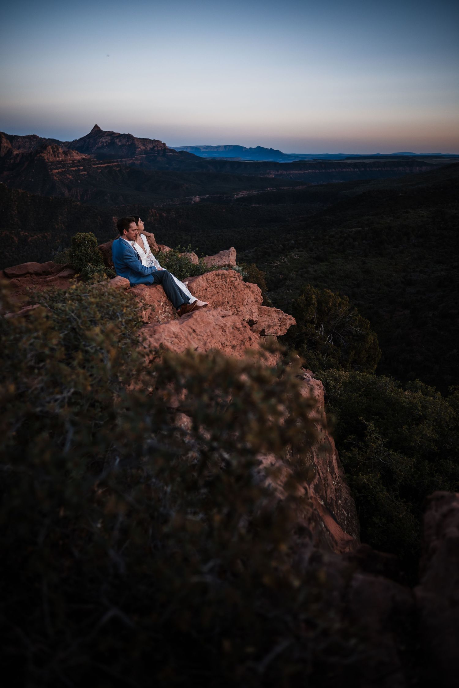 Groom in blue suit sitting alone on red rock cliff edge enjoying scenic desert sunset view.