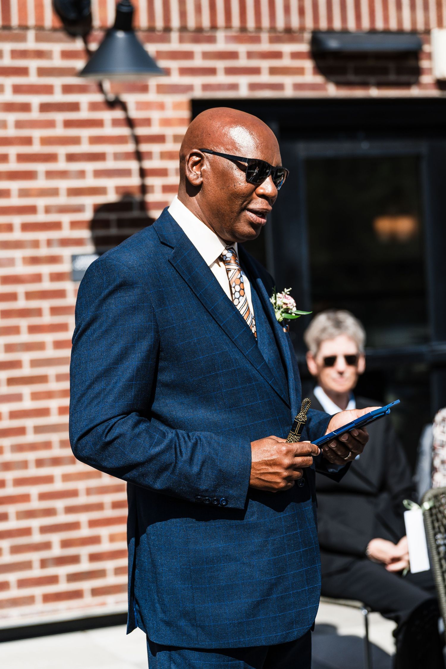Elegant man in blue suit with boutonniere reads from tablet at outdoor wedding ceremony.