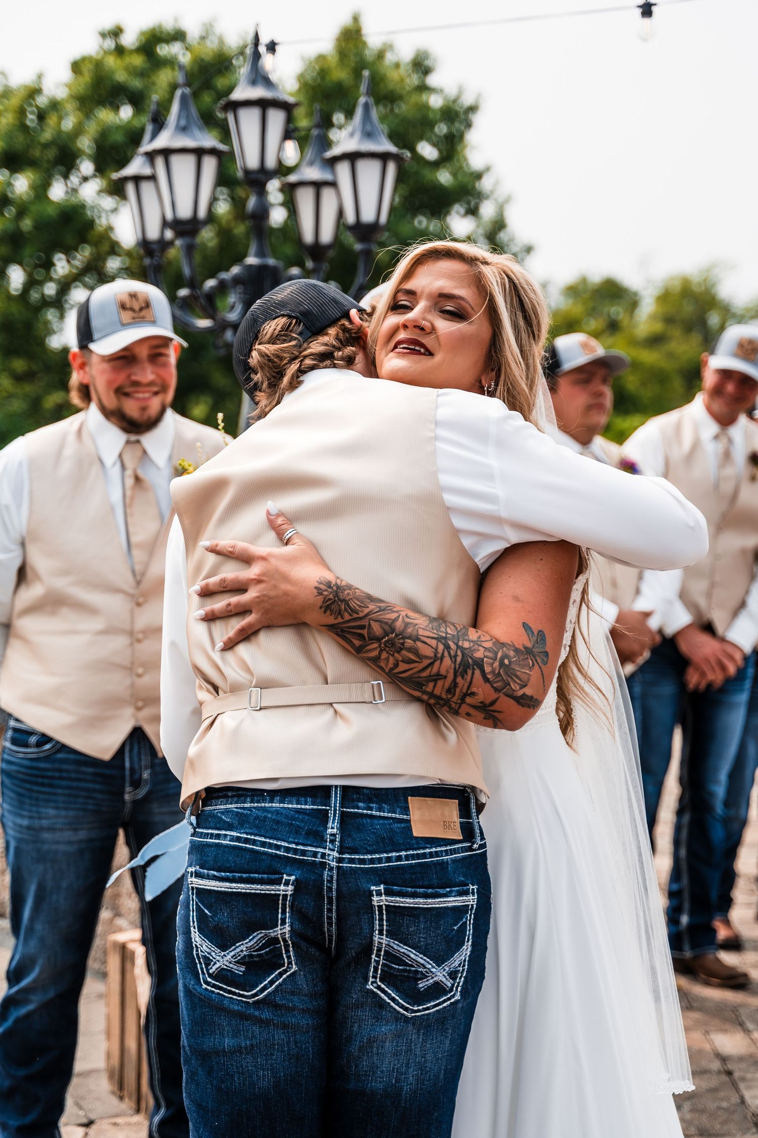 A tender embrace shared between a bride in white and guest with visible tattoos at an outdoor wedding celebration.