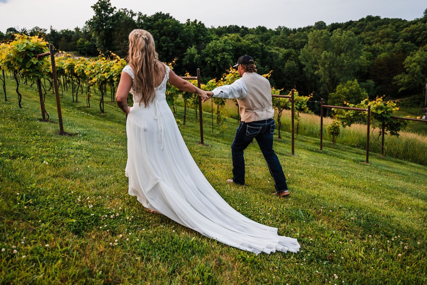 Wedding couple walking through vineyard with flowing white dress trailing behind.