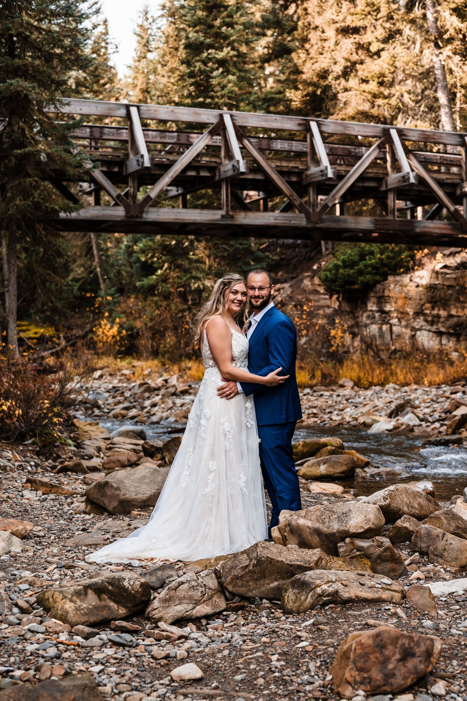 Wedding couple poses on rocky terrain near a rustic wooden bridge in autumn scenery.