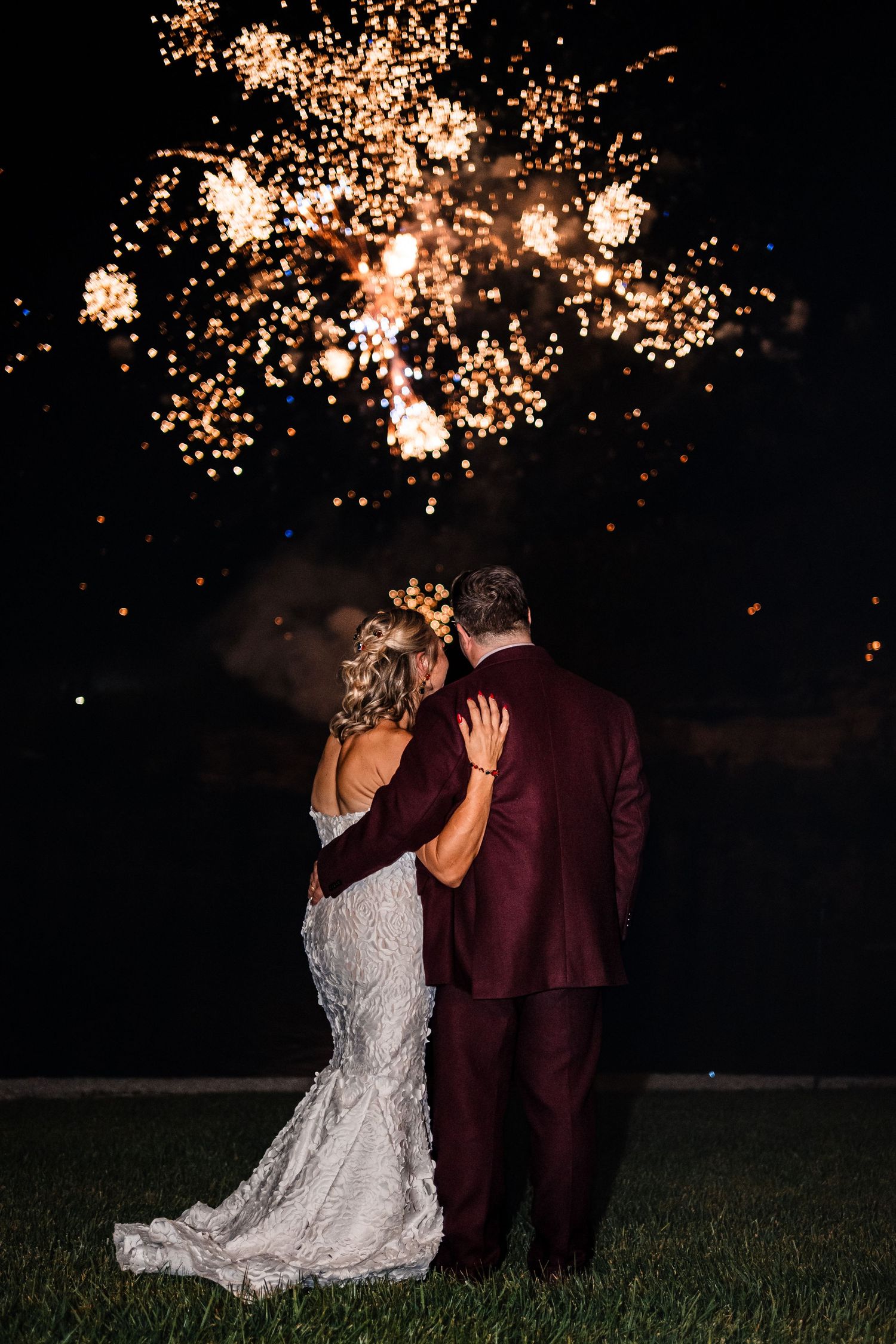 Couple watching fireworks display at night with bride in white dress and groom in burgundy suit.