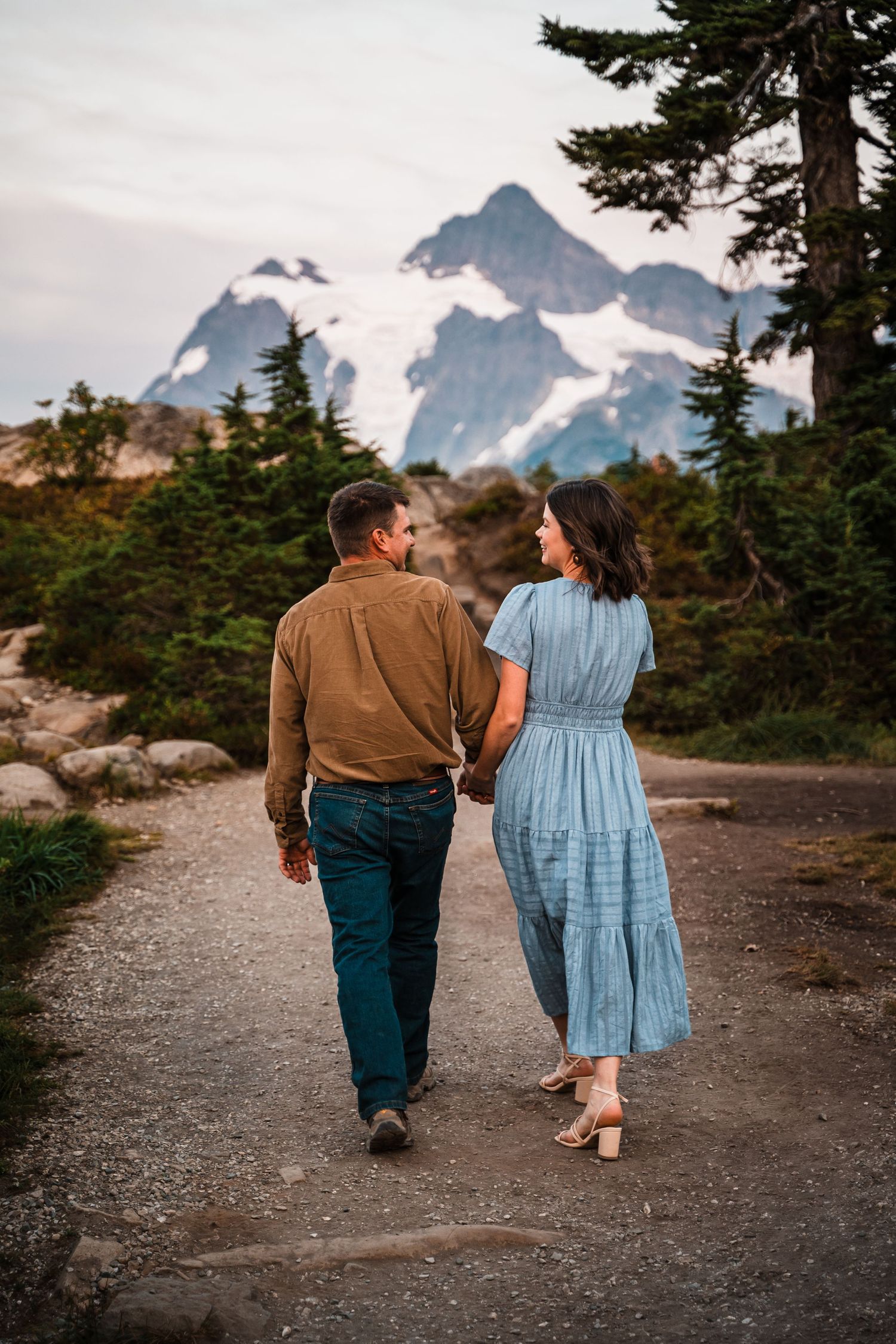 A couple walks hand in hand along a mountain trail with evergreen trees and snow-capped peaks in the distance.