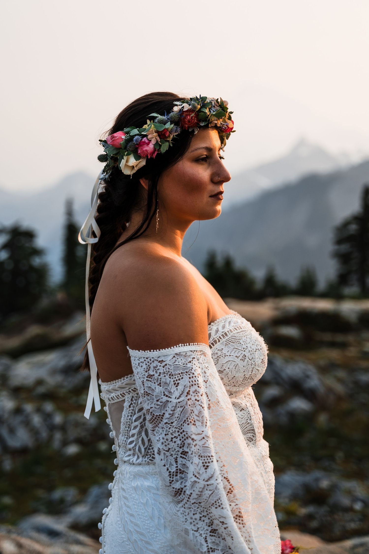 Bride in lace off-shoulder dress with floral crown against mountain backdrop at sunset.