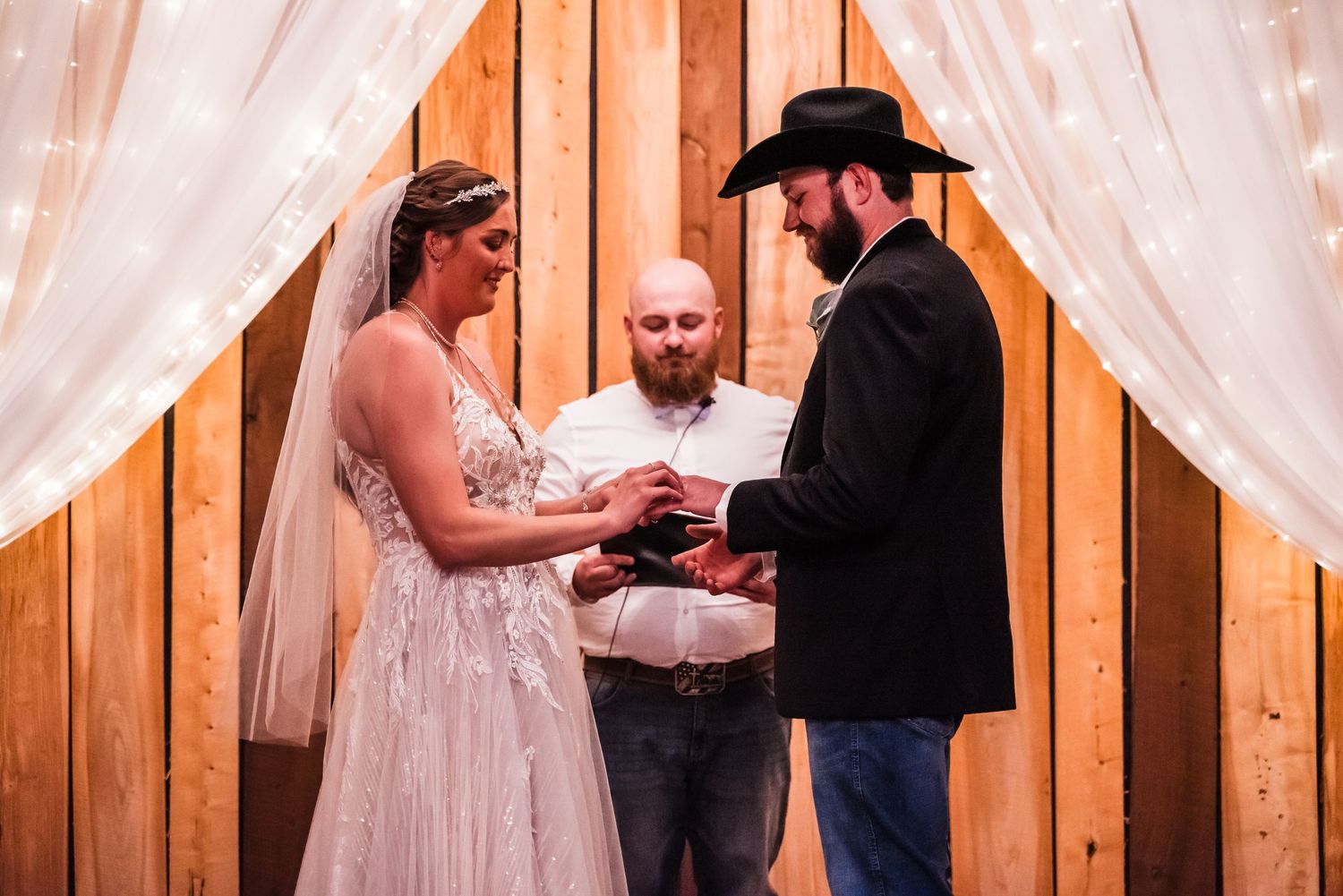 Wedding ceremony in front of wooden wall with white curtains as couple exchanges rings with officiant.