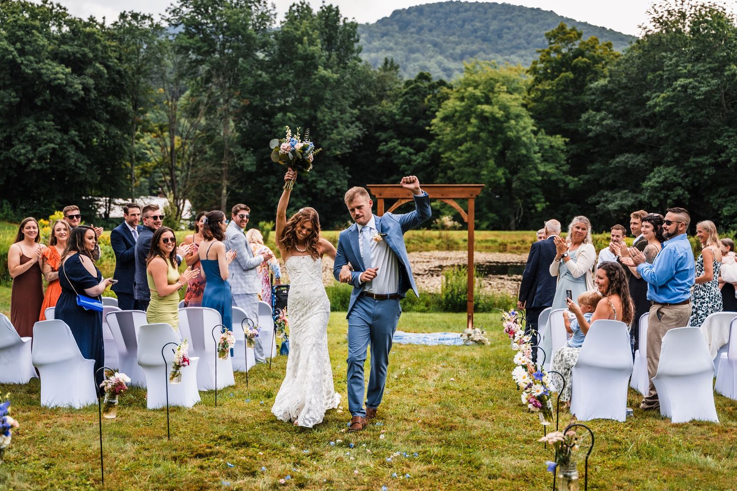 A joyful outdoor wedding ceremony with guests seated on white chairs and mountains in the background.