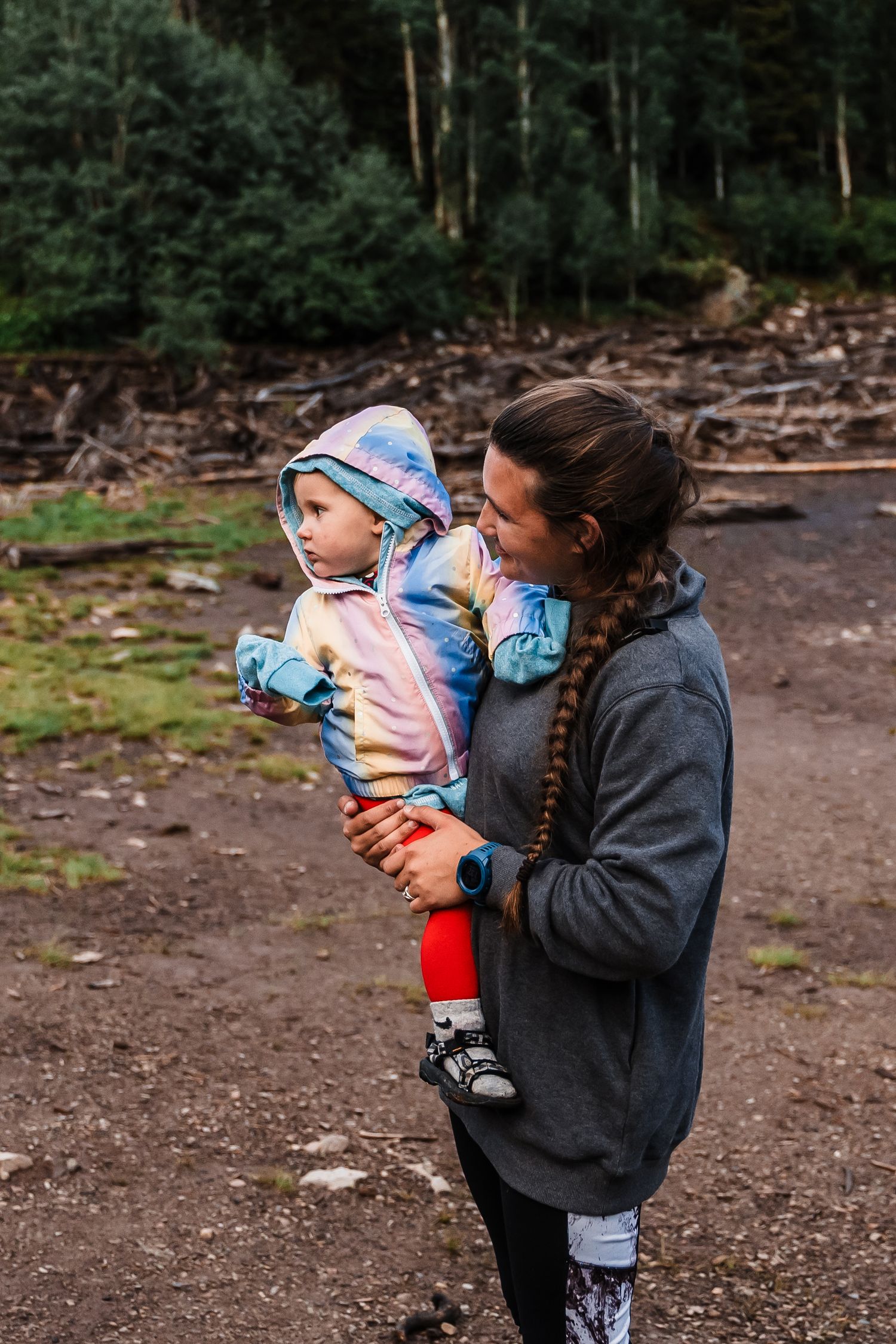 A parent and child in warm clothing share a tender moment on a forest trail surrounded by fallen logs and evergreen trees.