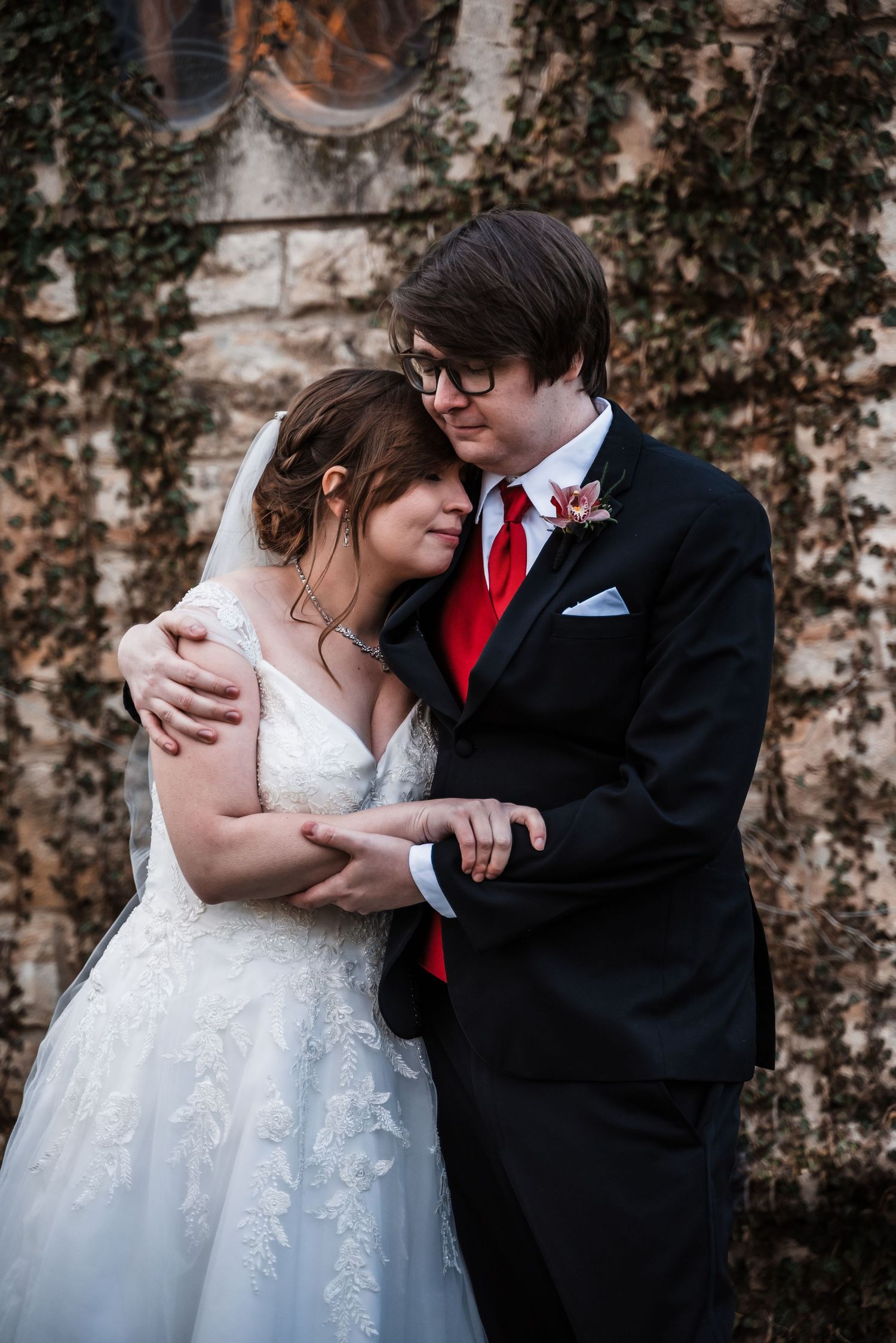 A romantic wedding couple embraces in front of an ivy-covered stone church wall on their winter wedding day.