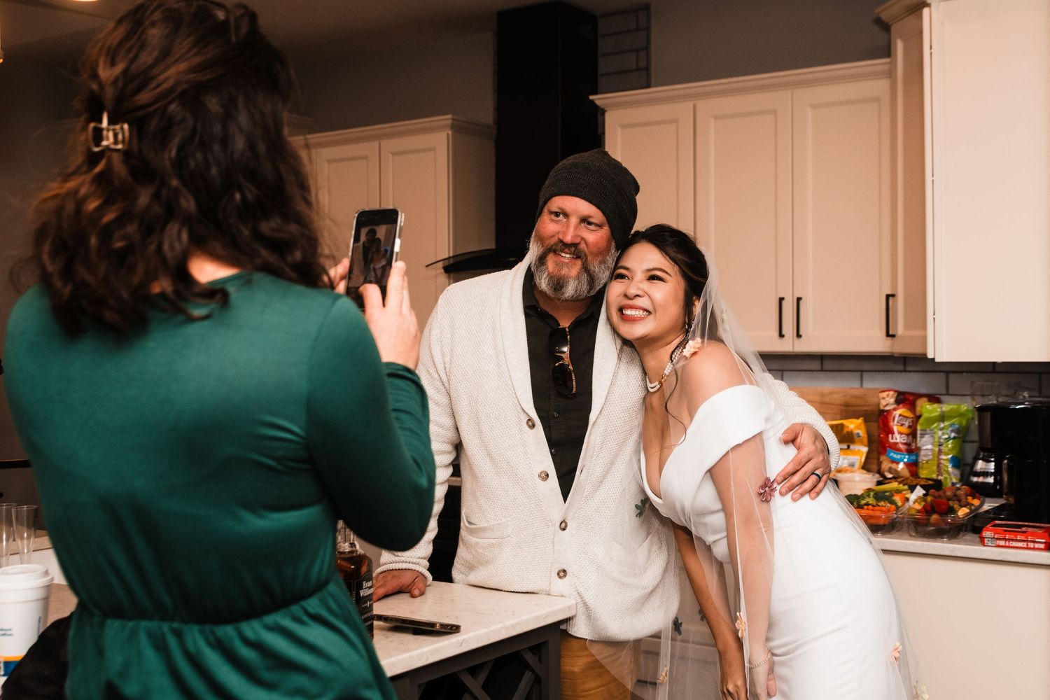 A couple poses in a kitchen while someone in a green dress takes their photo on a phone.