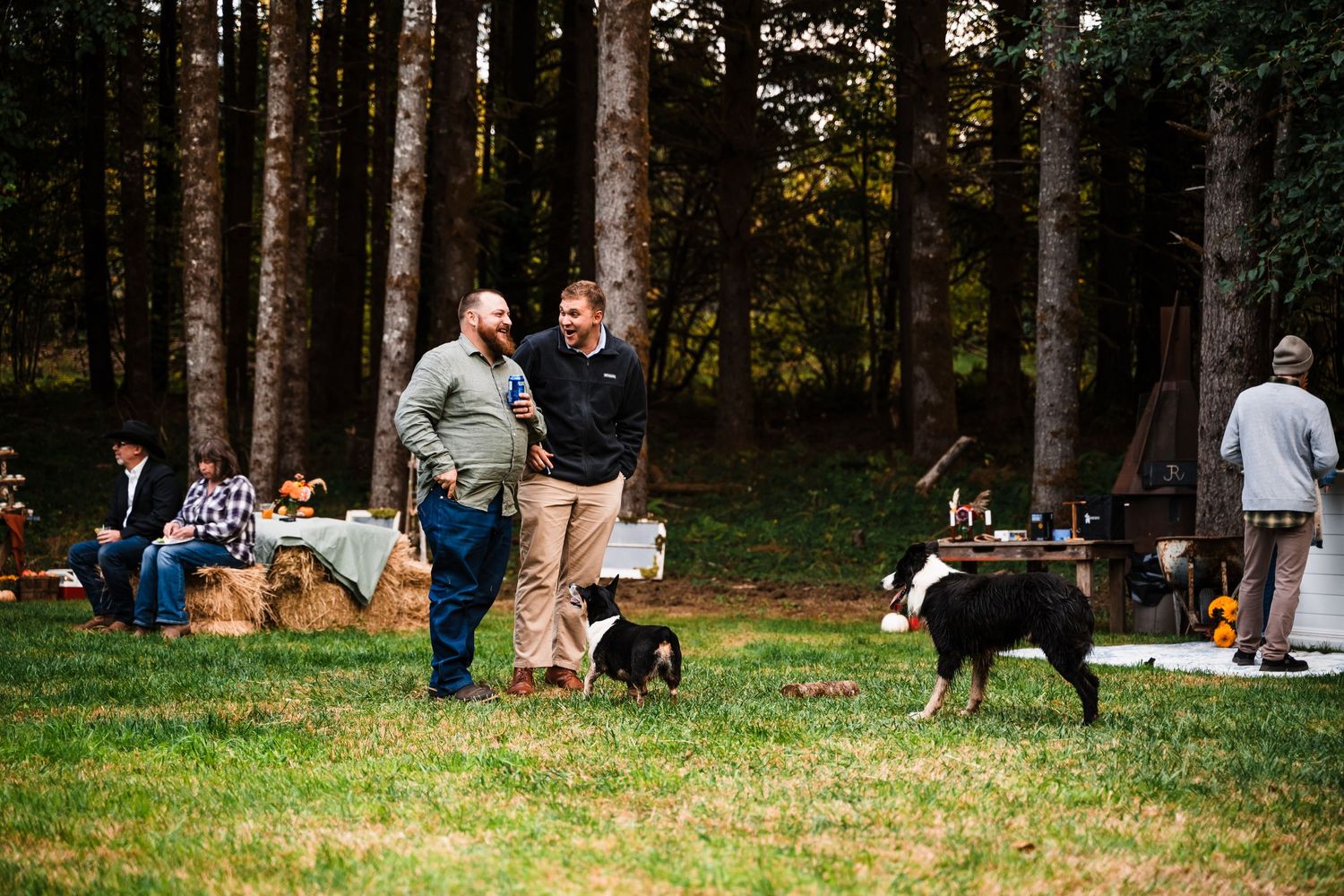 Friends and dogs enjoying a casual outdoor gathering on a grassy lawn near woods.