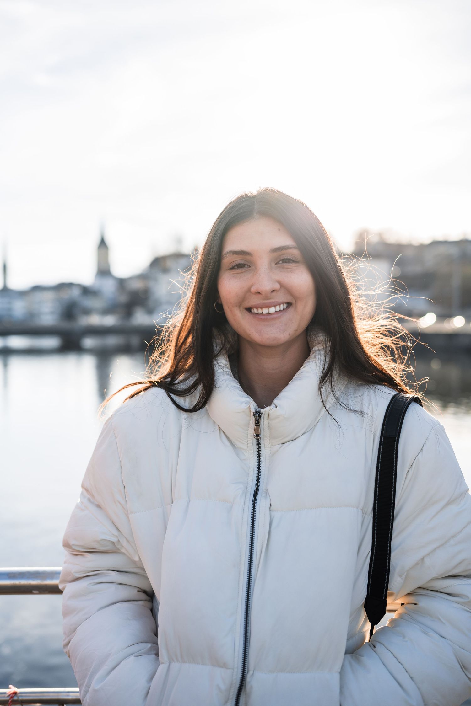 Person wearing a white winter coat poses by a waterfront marina at sunset.