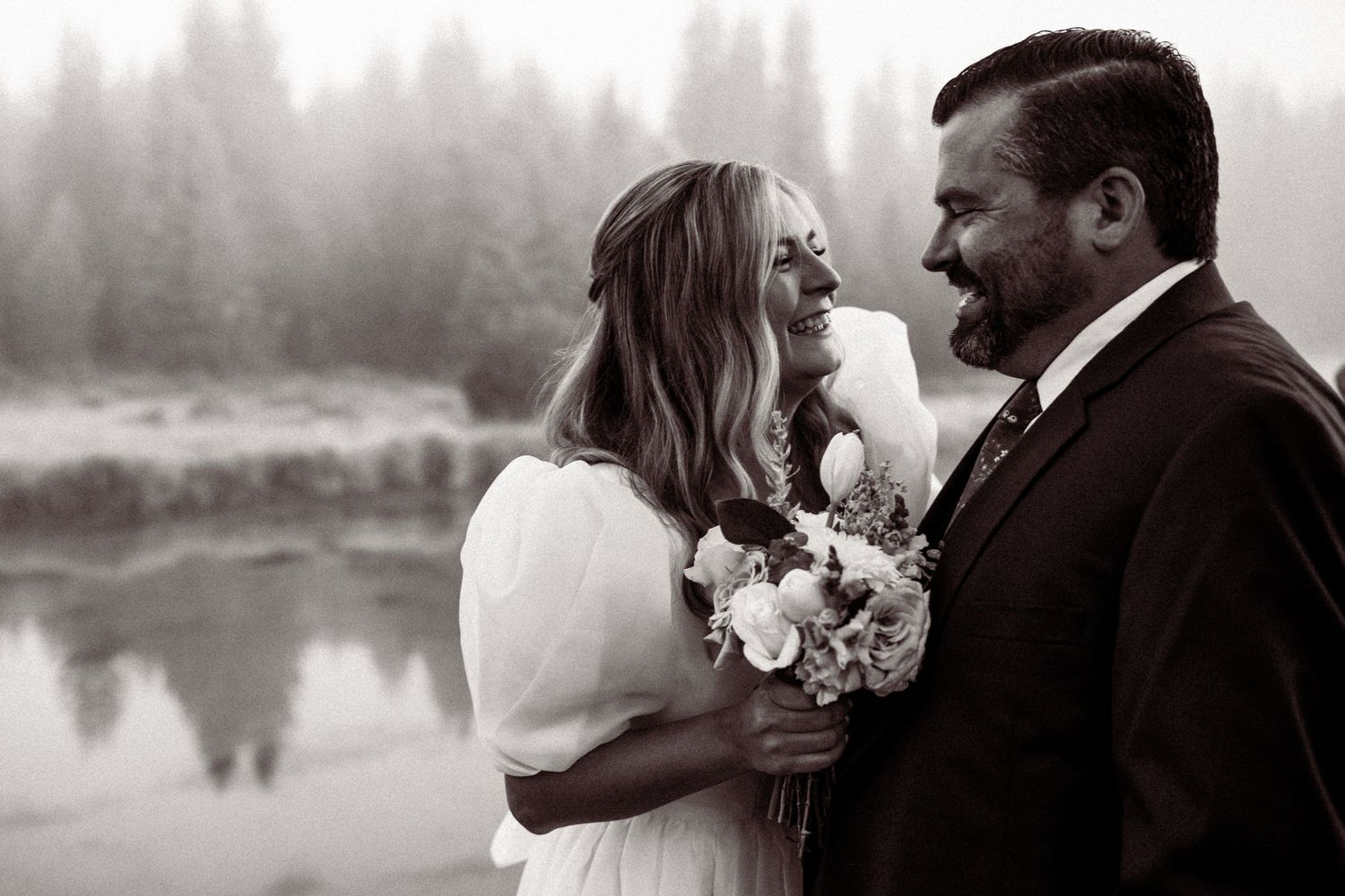 Black and white wedding photo of a couple sharing a joyful moment by a misty lake while holding a floral bouquet.