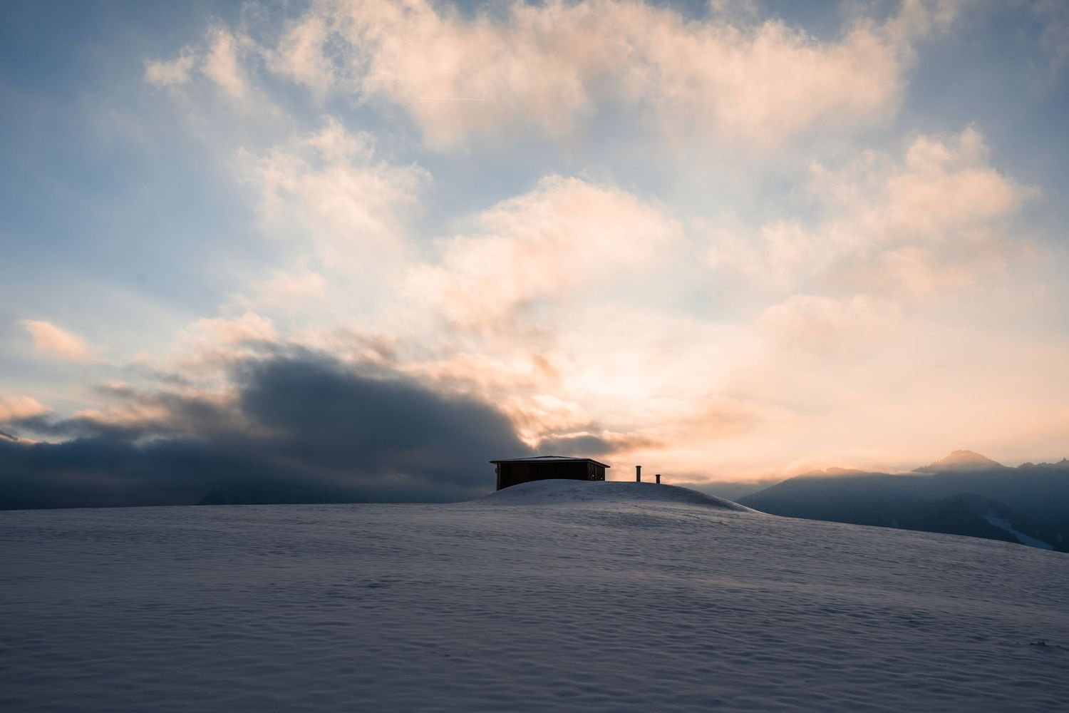 A panoramic view of a cloudy sunset sky over snow-covered mountains with silhouetted peaks.