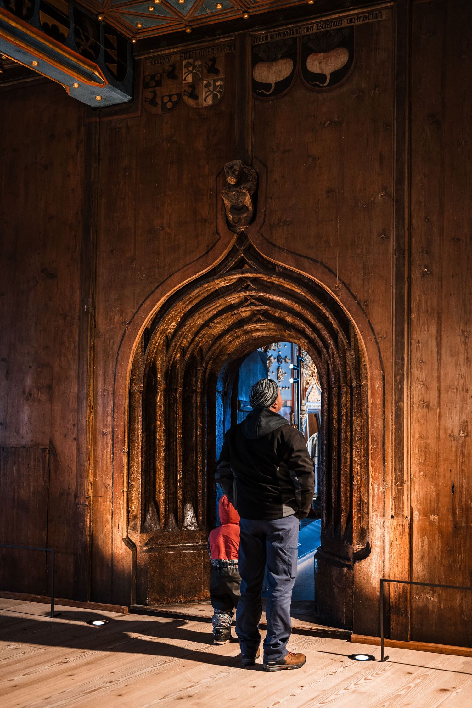 People walk through a dimly lit Gothic archway corridor with wooden panels and vaulted ceilings.