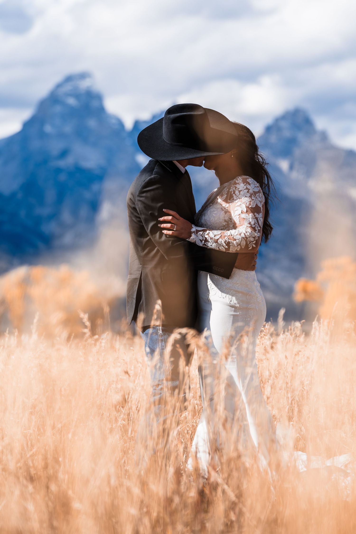 A couple shares an intimate moment in a golden wheat field with majestic mountain peaks rising in the background.