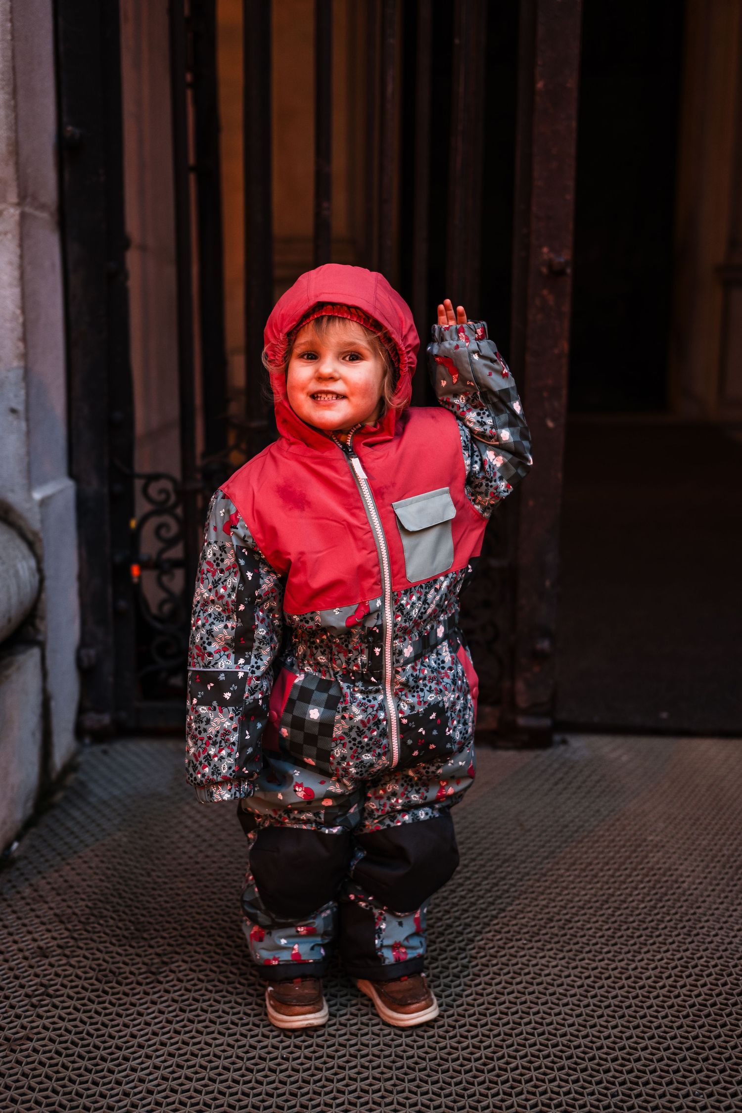 A small child in a red hooded coat and floral print outfit crouches on a textured metal floor.
