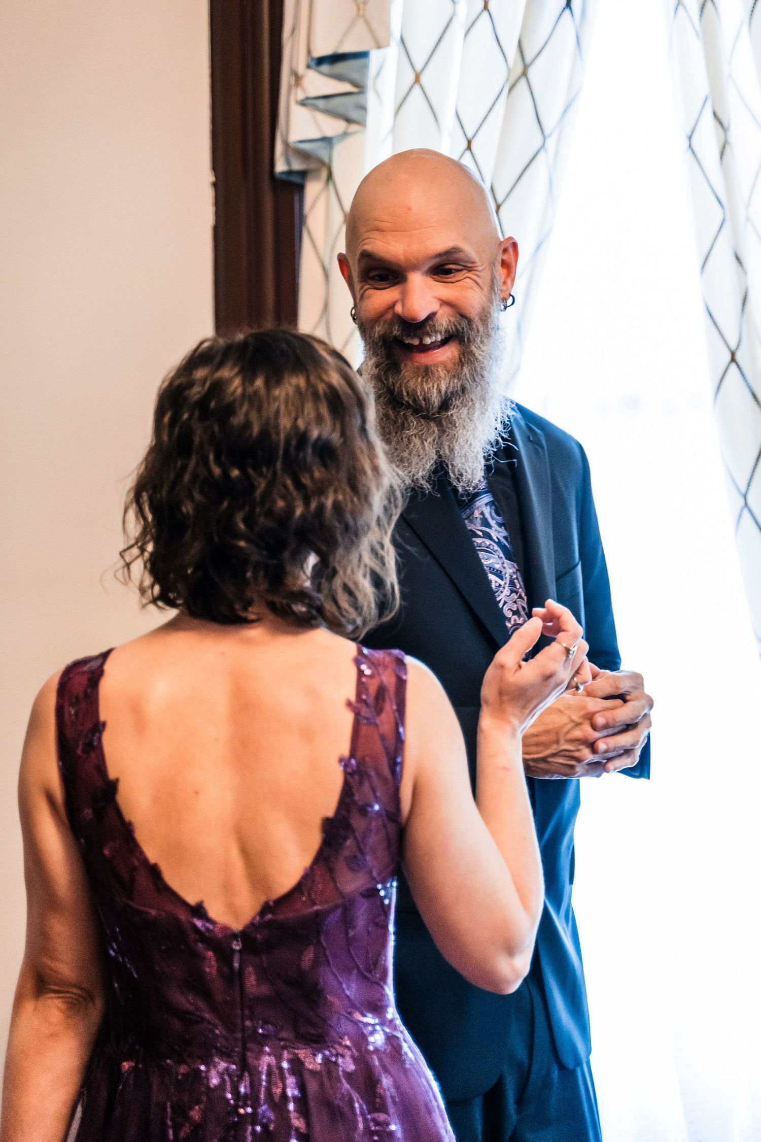 Bald groom with gray beard smiling at bride in purple sequin dress during wedding ceremony.