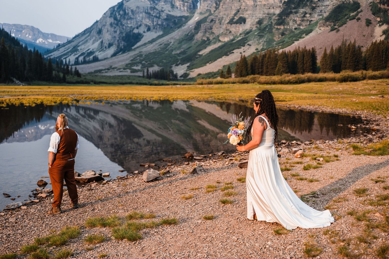 Bride and groom walking near reflective alpine lake with mountain backdrop at sunset elopement.