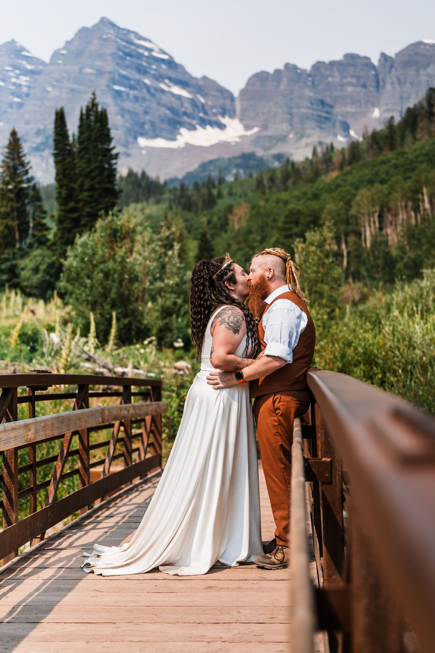 Couple kissing on wooden bridge with Maroon Bells mountain peaks and green forest behind them.