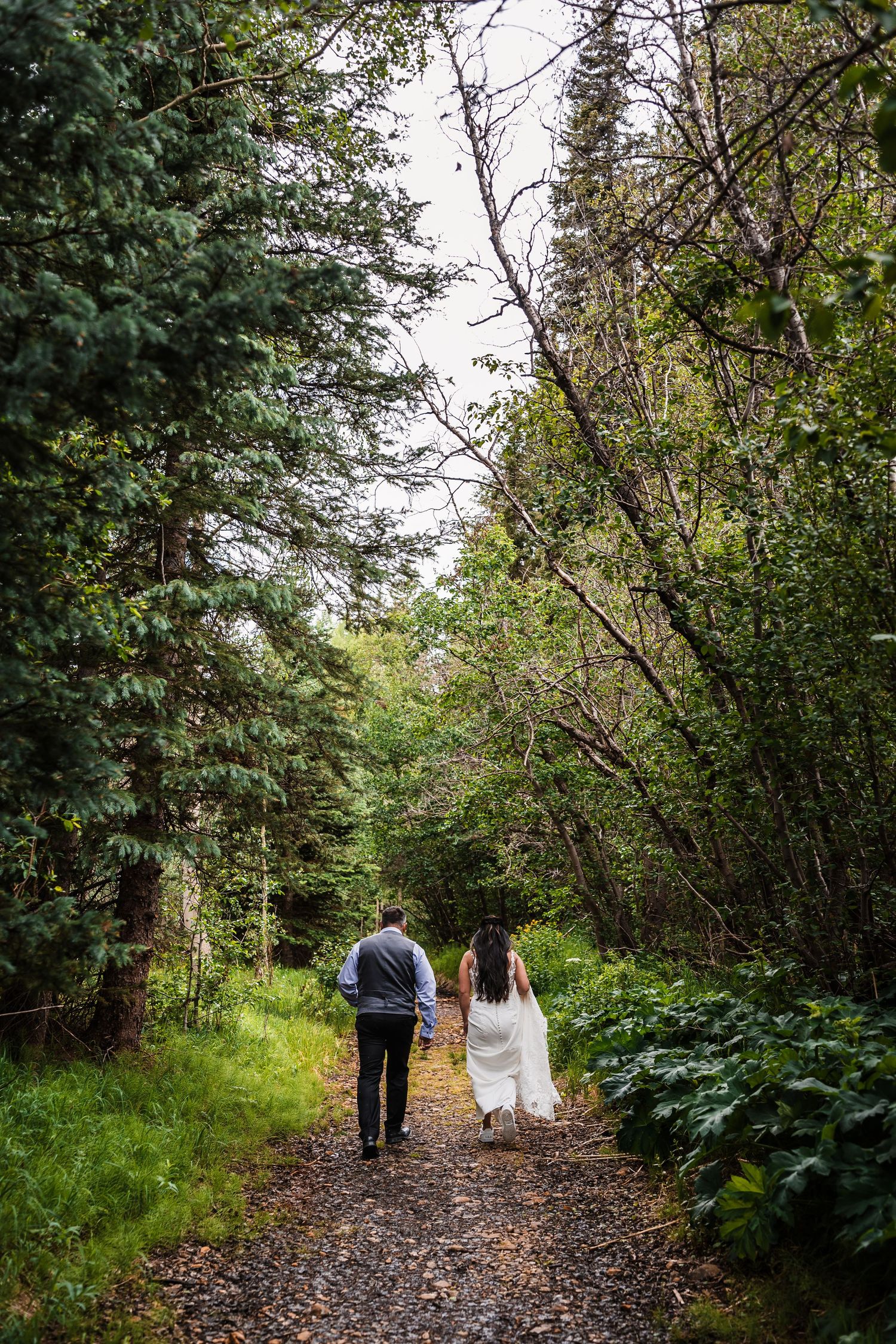 Bride and groom walk hand in hand down a lush forest trail during their mountain elopement adventure.