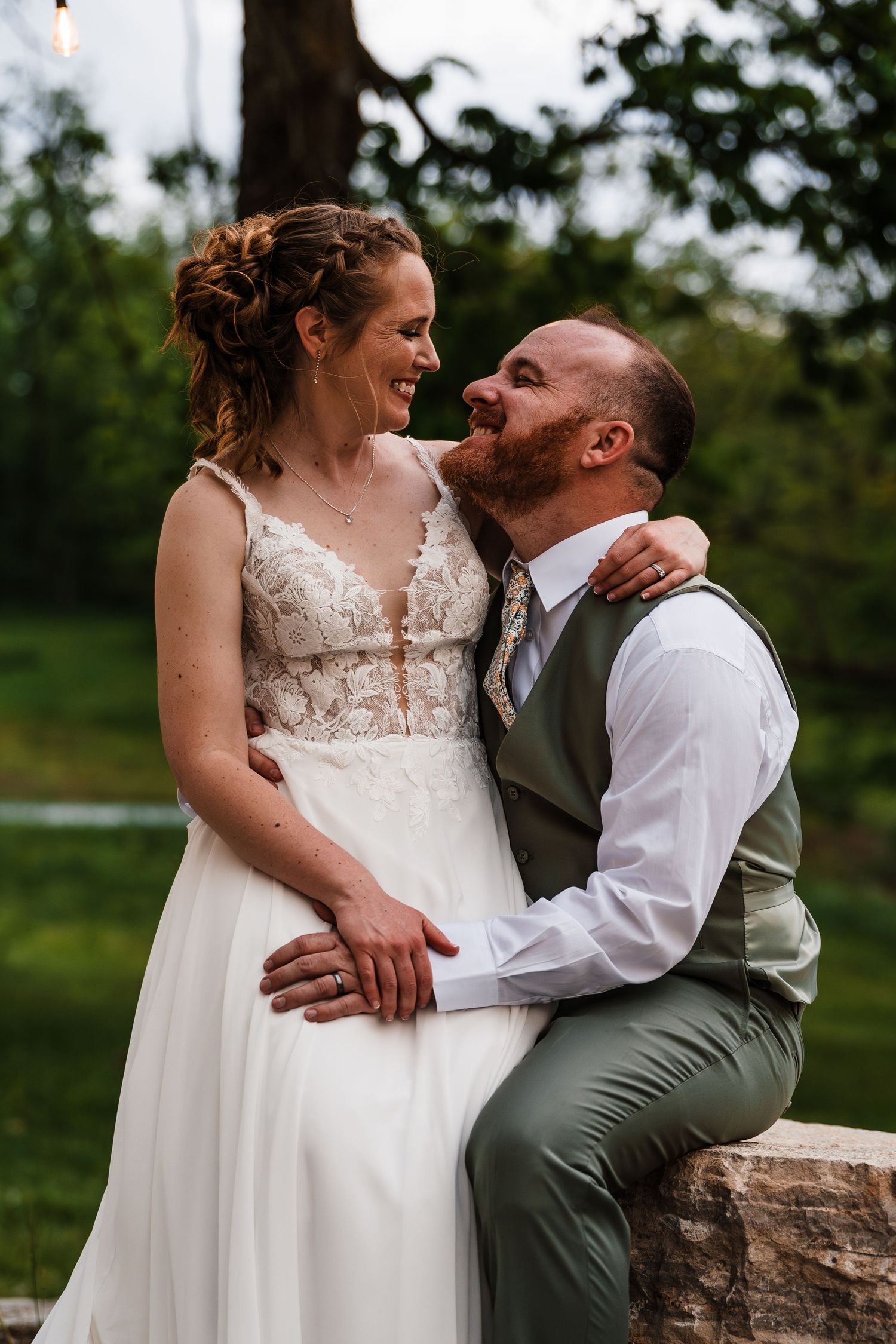 Bride and groom share joyful intimate moment outdoors on stone wall at sunset.