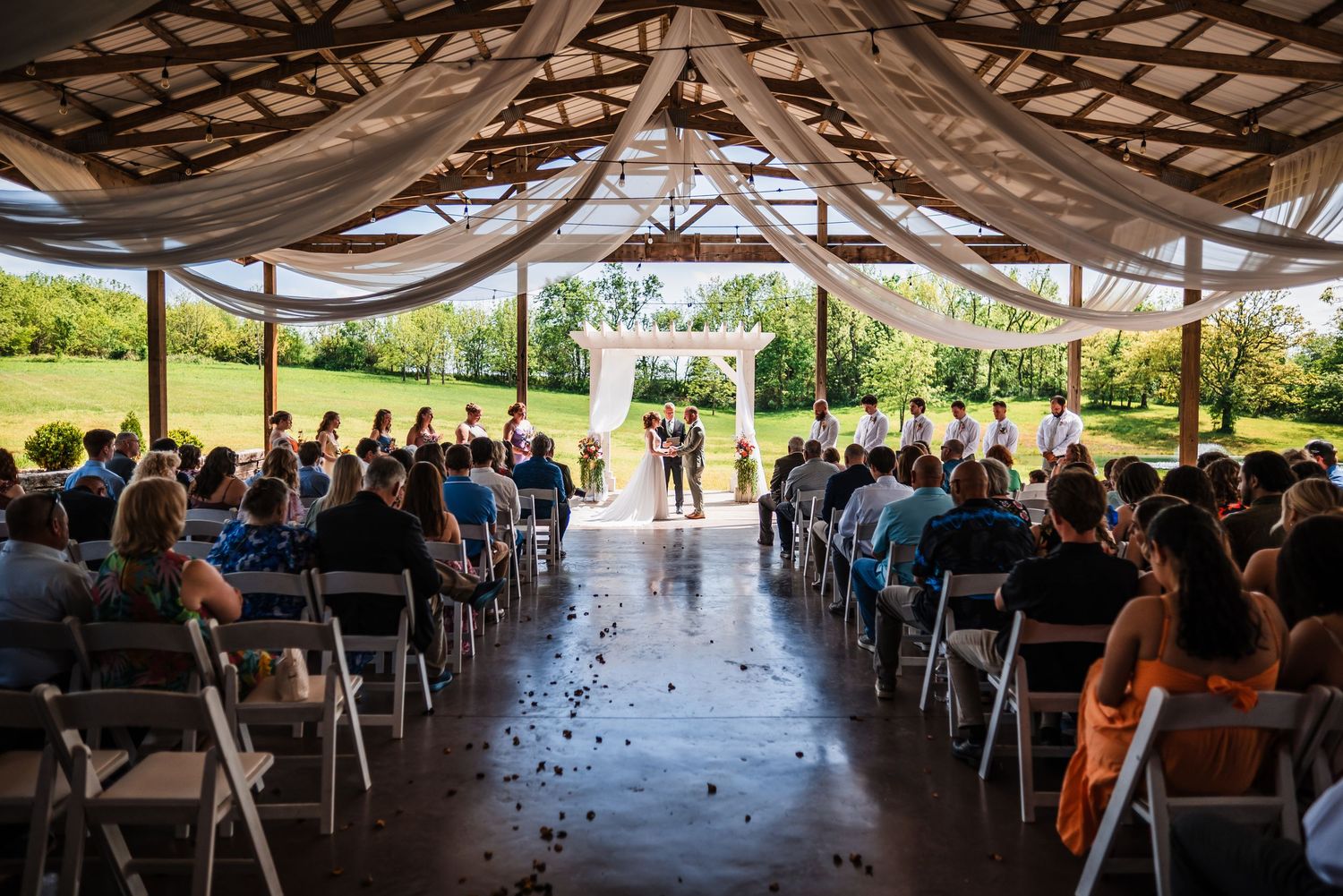 Elegant outdoor wedding ceremony under a draped pavilion with guests seated in white chairs along a petal-lined aisle.