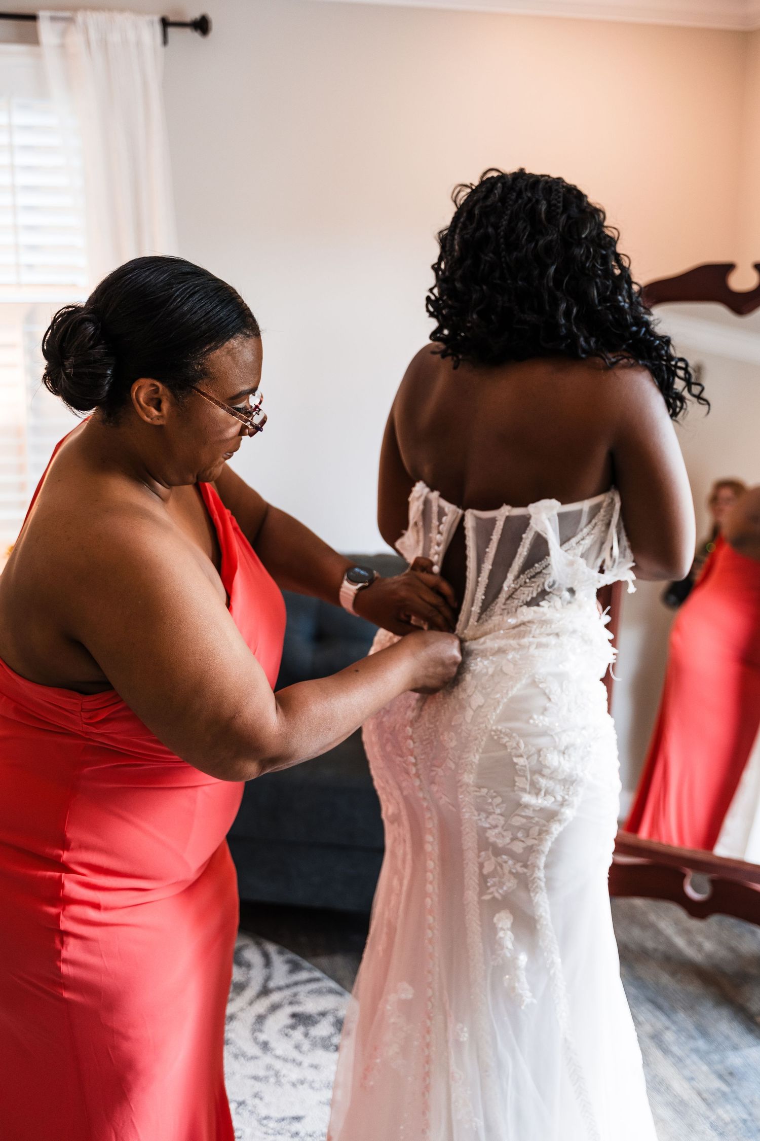Woman in red dress lacing up a bride in a white lace wedding gown from behind in a bright room.
