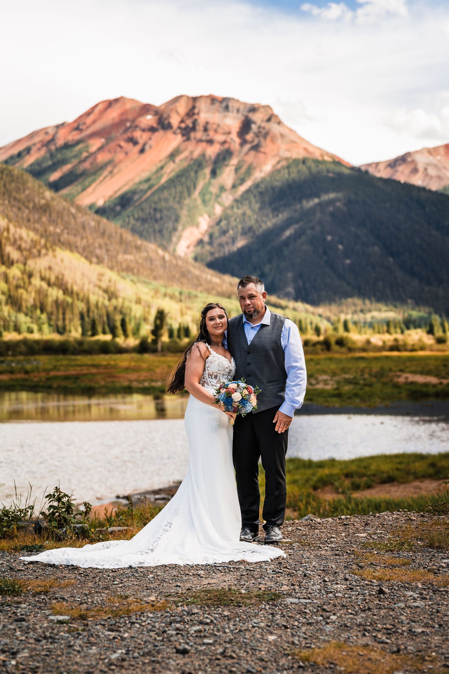 Bride and groom posing with bouquet before a stunning red mountain and alpine lake backdrop.