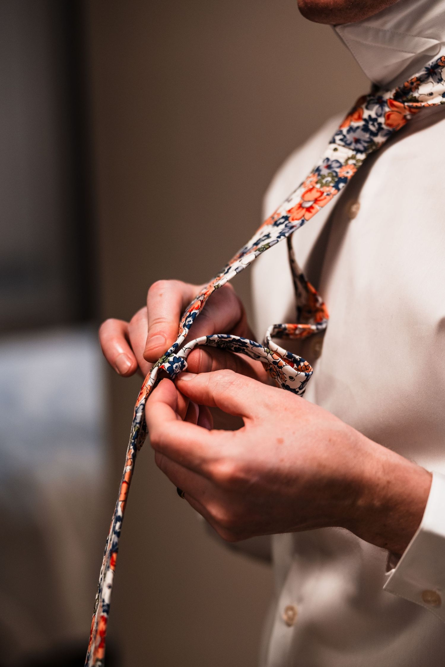 Close-up of groom tying a floral patterned necktie while wearing a white dress shirt.