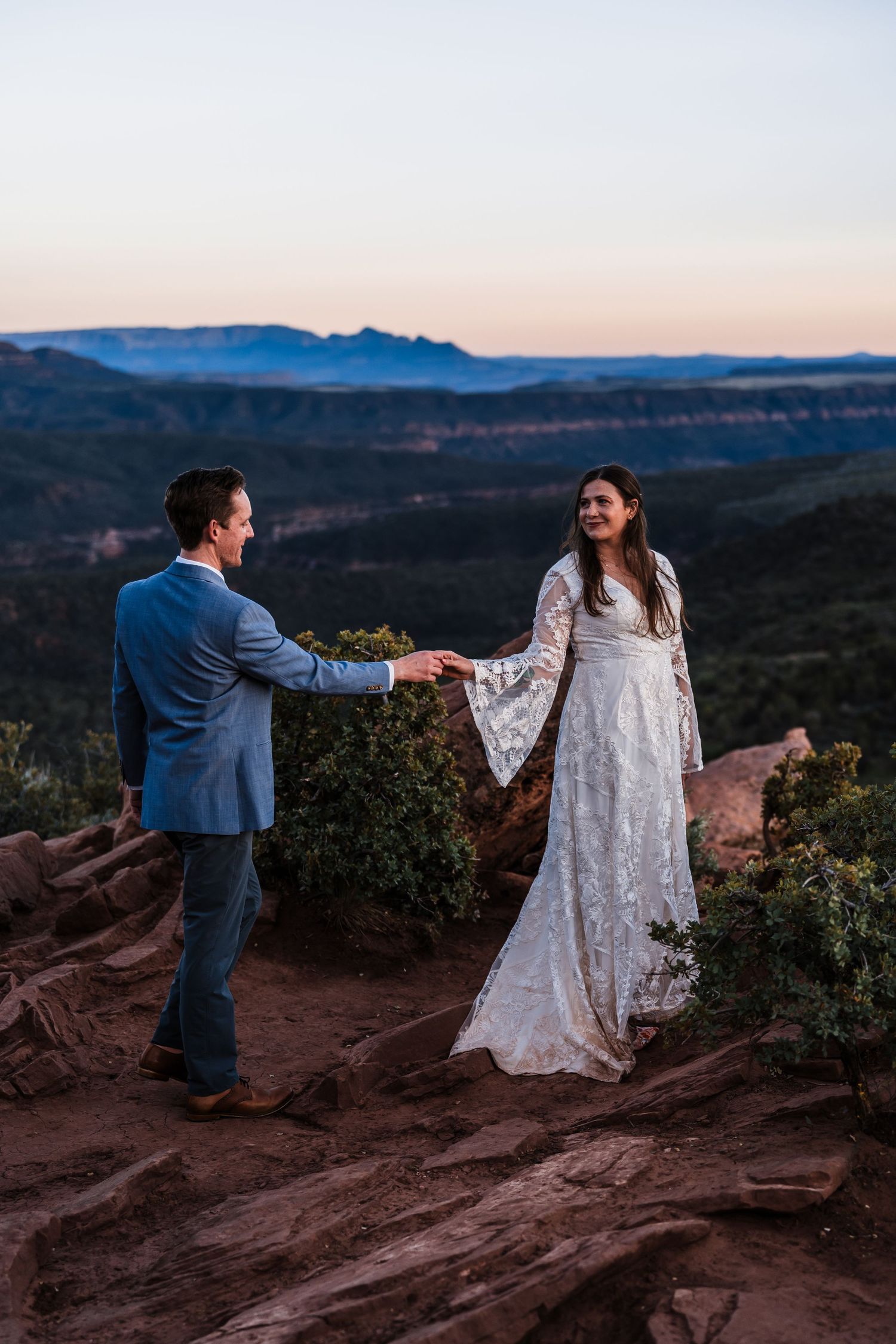 Bride in lace gown and groom in blue suit holding hands on red rock overlook at dusk.