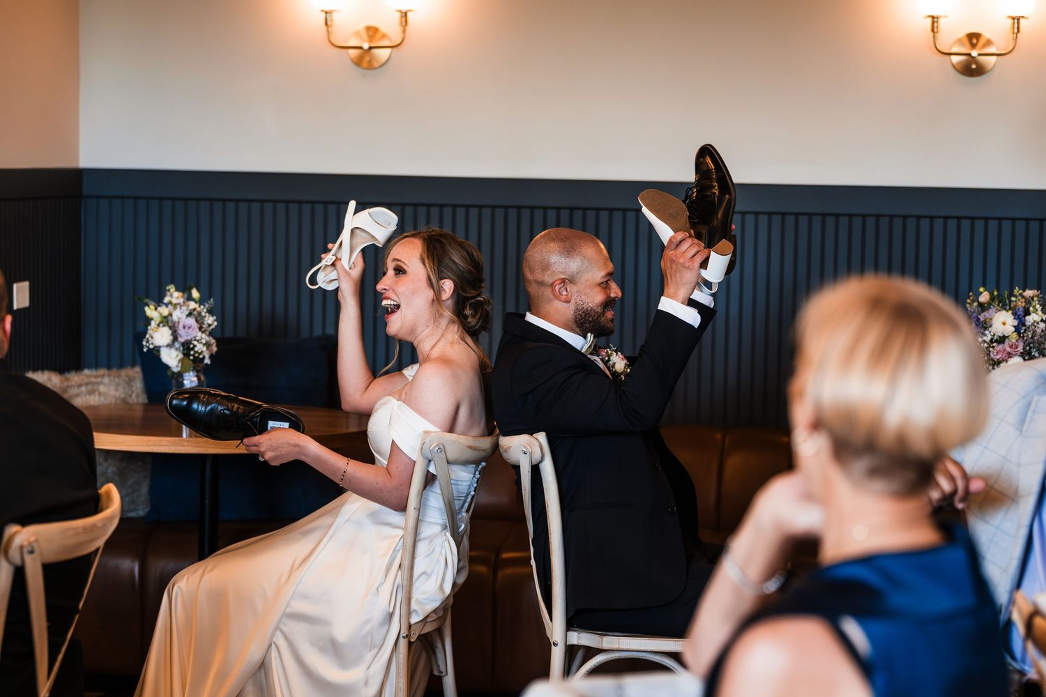 Bride and groom laughing and holding up shoes during fun wedding reception shoe game activity.