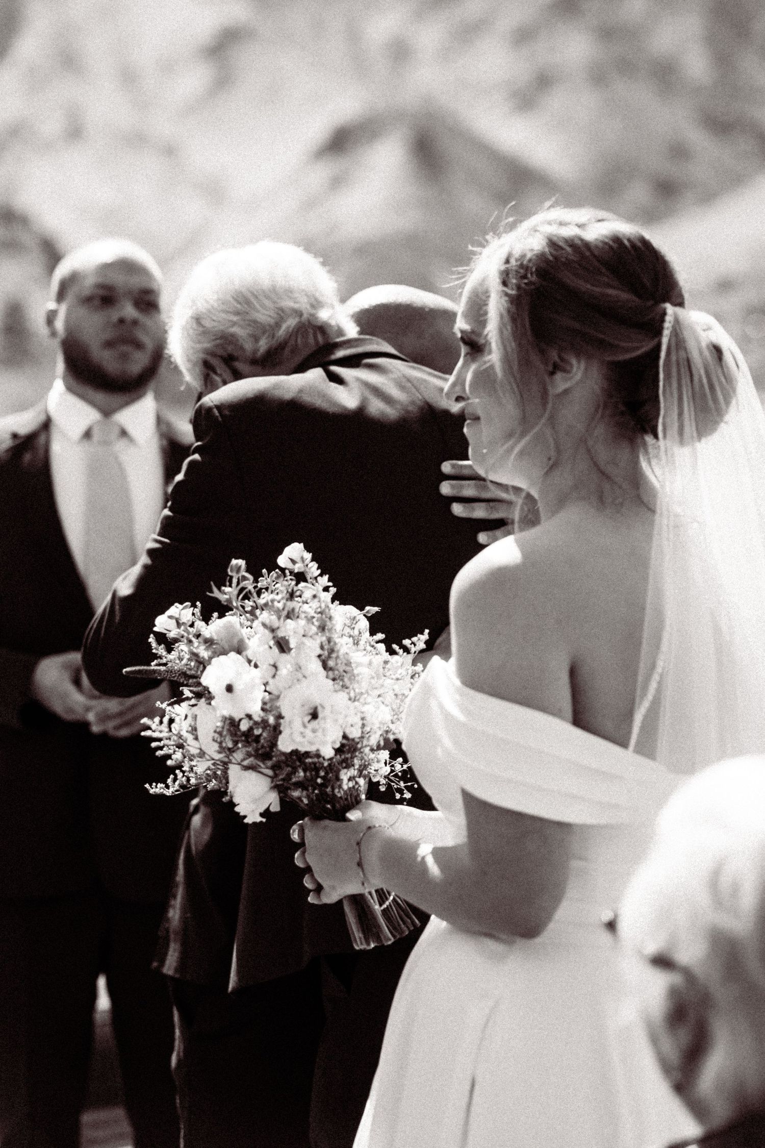 Black and white photo of bride holding bouquet watching an embrace during outdoor wedding ceremony.